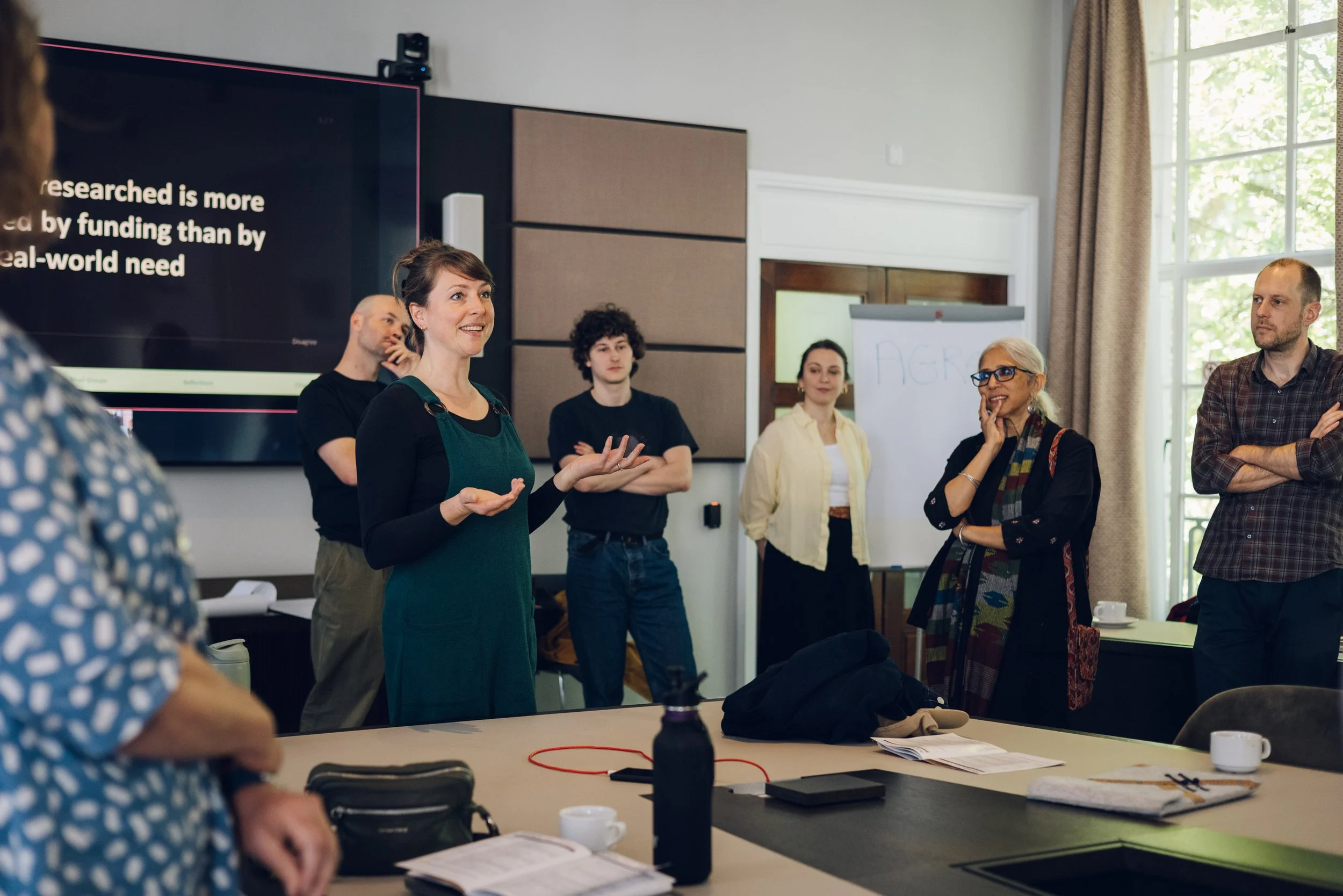 A woman is speaking to a group of diverse people in a conference room with large windows. A large screen behind her shows partially visible text. The audience appears attentive, with some people smiling and others listening thoughtfully.