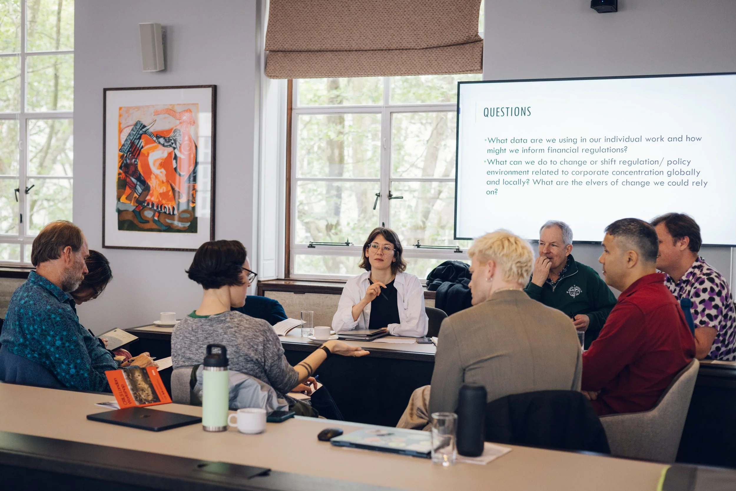 A group of people attending a meeting or conference in a room with large windows. A woman is seated at a table with notebooks and cups, and a large screen displays questions related to data and regulatory environment.