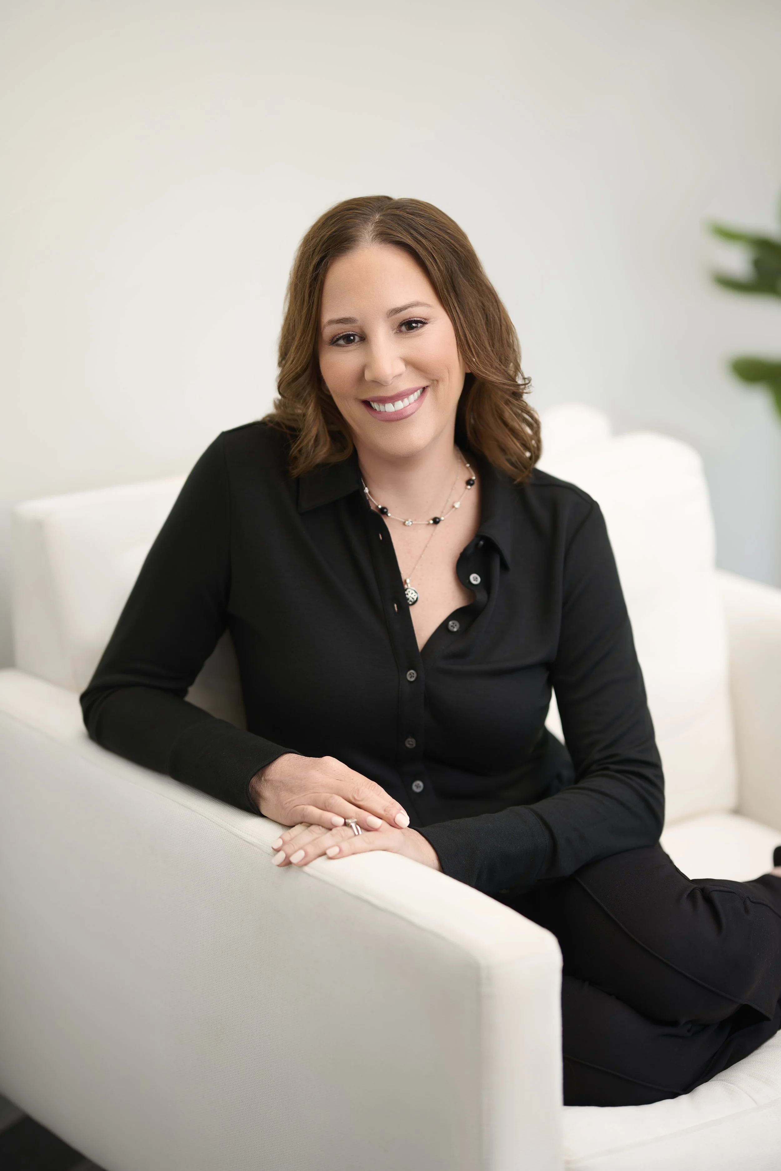 A smiling woman with shoulder-length brown hair, wearing a black button-up shirt and jewelry, sitting on a white sofa in a bright room.