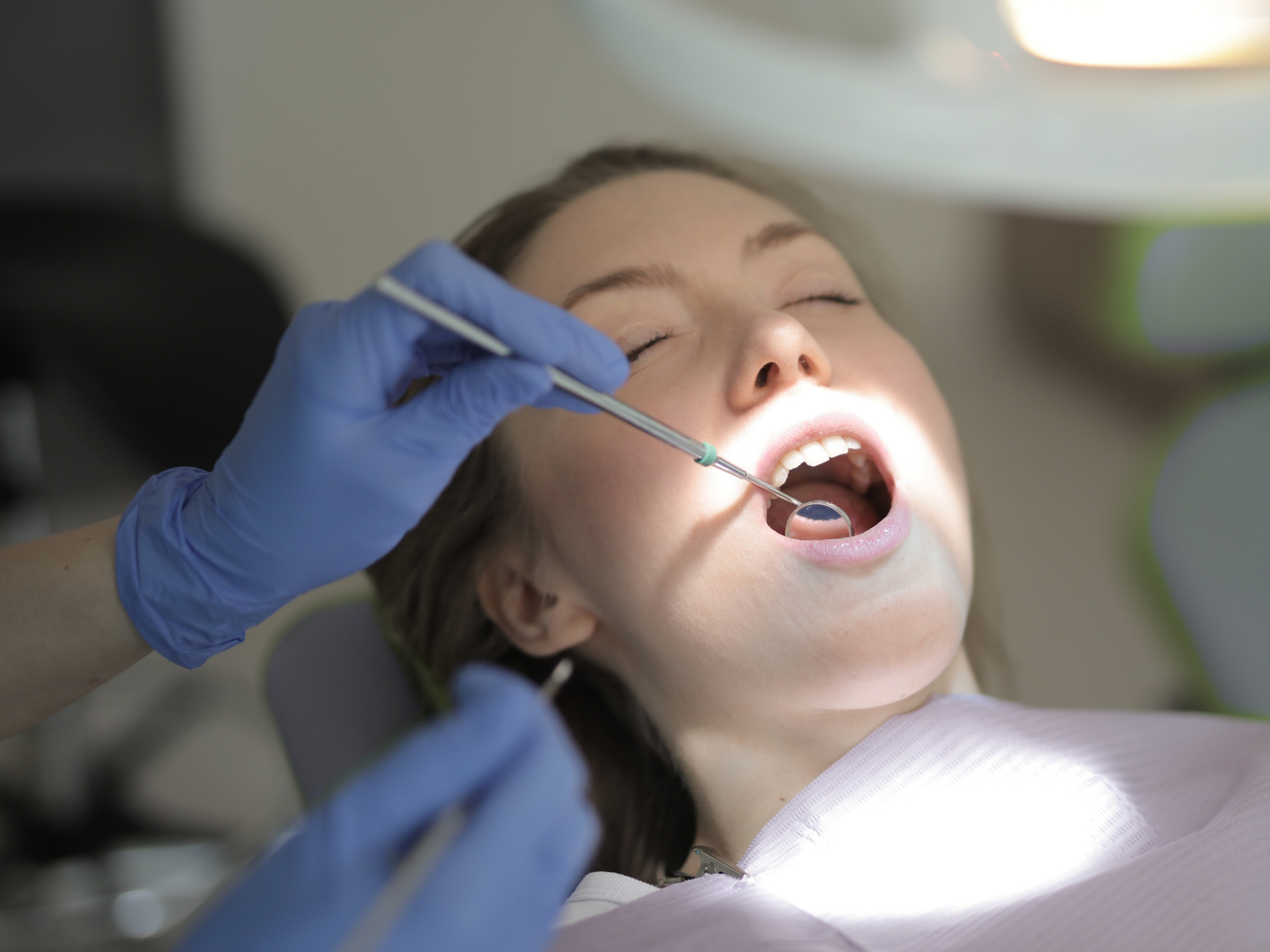 Person receiving dental treatment, lying in a dental chair with a dental mirror in their mouth, while a dentist in blue gloves uses a dental tool.