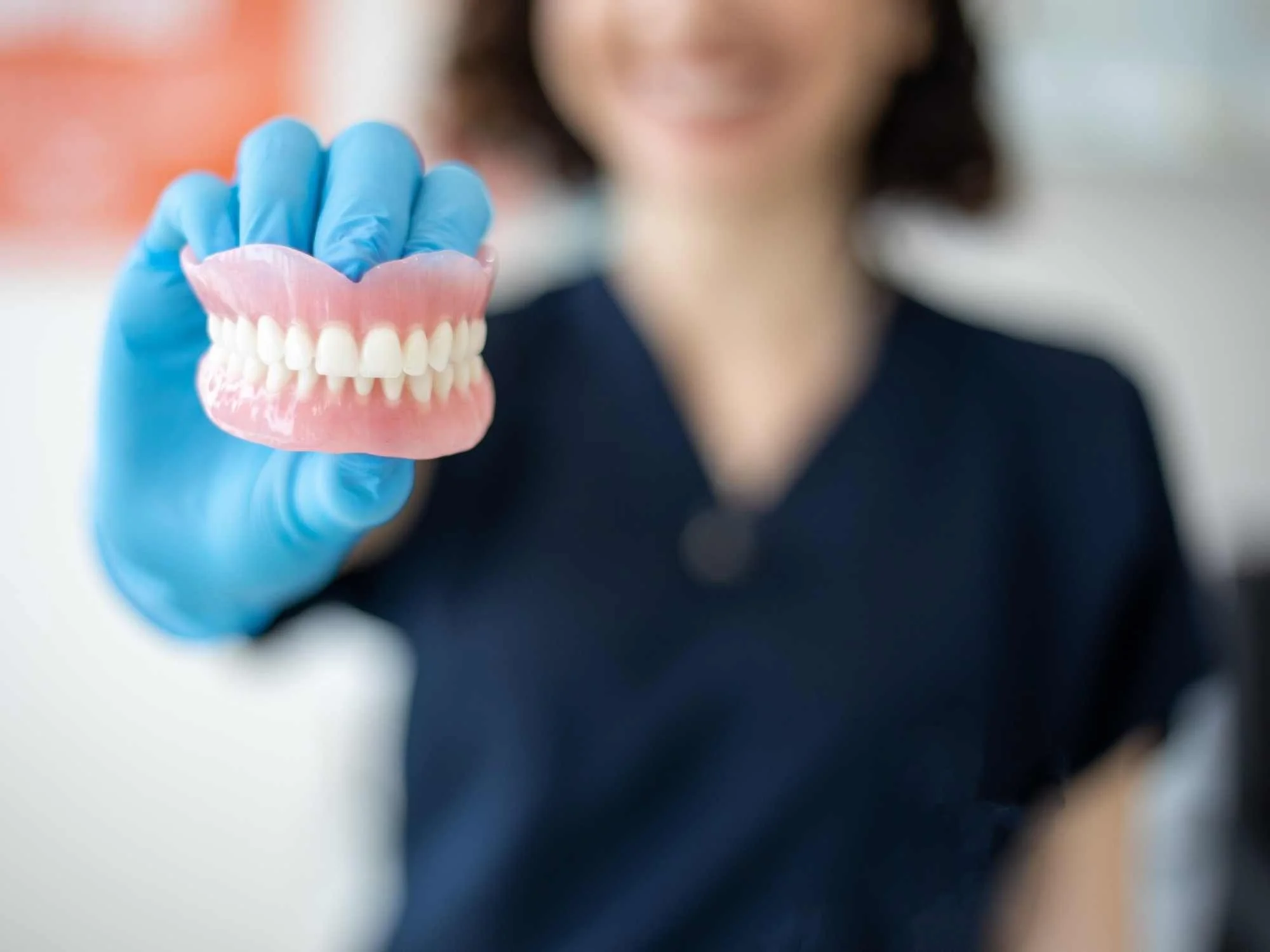 A woman wearing blue gloves holding a dental model with dentures, smiling in the background.
