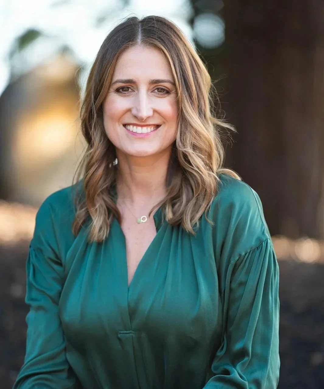 A woman with wavy, shoulder-length brown hair smiling outdoors, wearing a green blouse and a gold necklace.