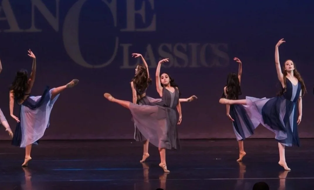 A group of dancers performing on stage in flowing dresses, with a backdrop displaying the words 'California Dance Classics'.