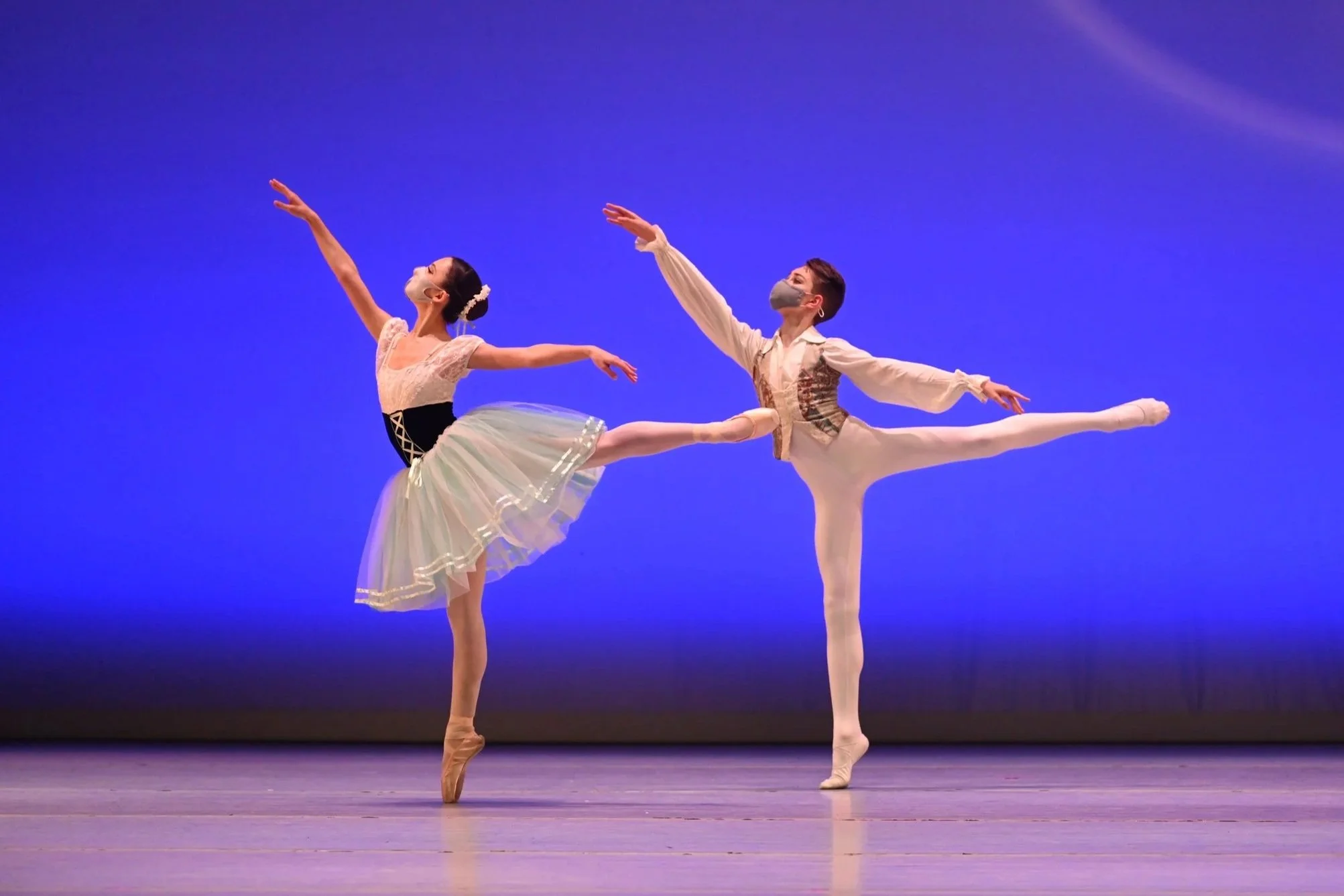 Ballet dancers performing on stage with a blue background, both wearing costumes and masks, with one dancer en pointe and the other in a pose with arms extended.