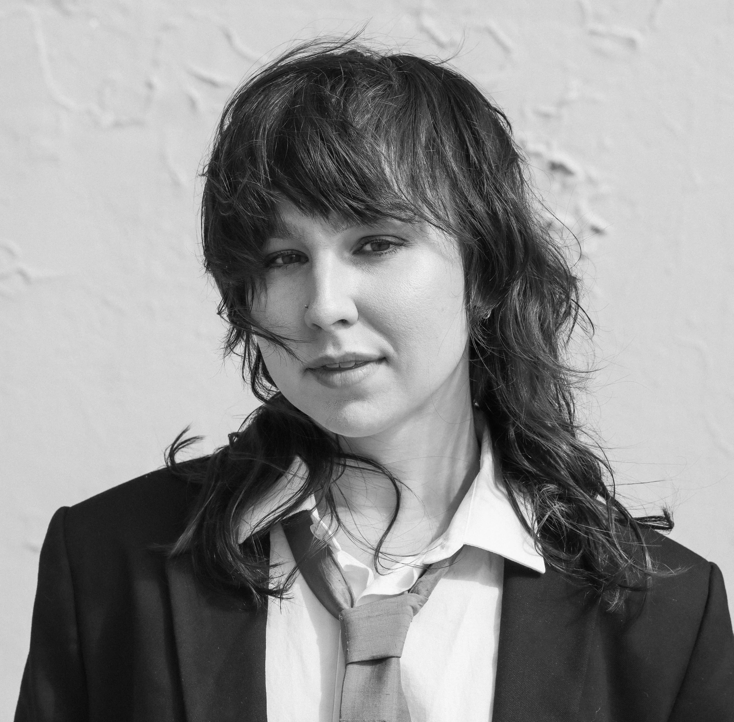 A woman with short, wavy brown hair, wearing makeup with silvery eyeshadow, hoop earrings, and a septum piercing, dressed in a sheer white lace top with polka dots and ruffles, posing against a dark background.