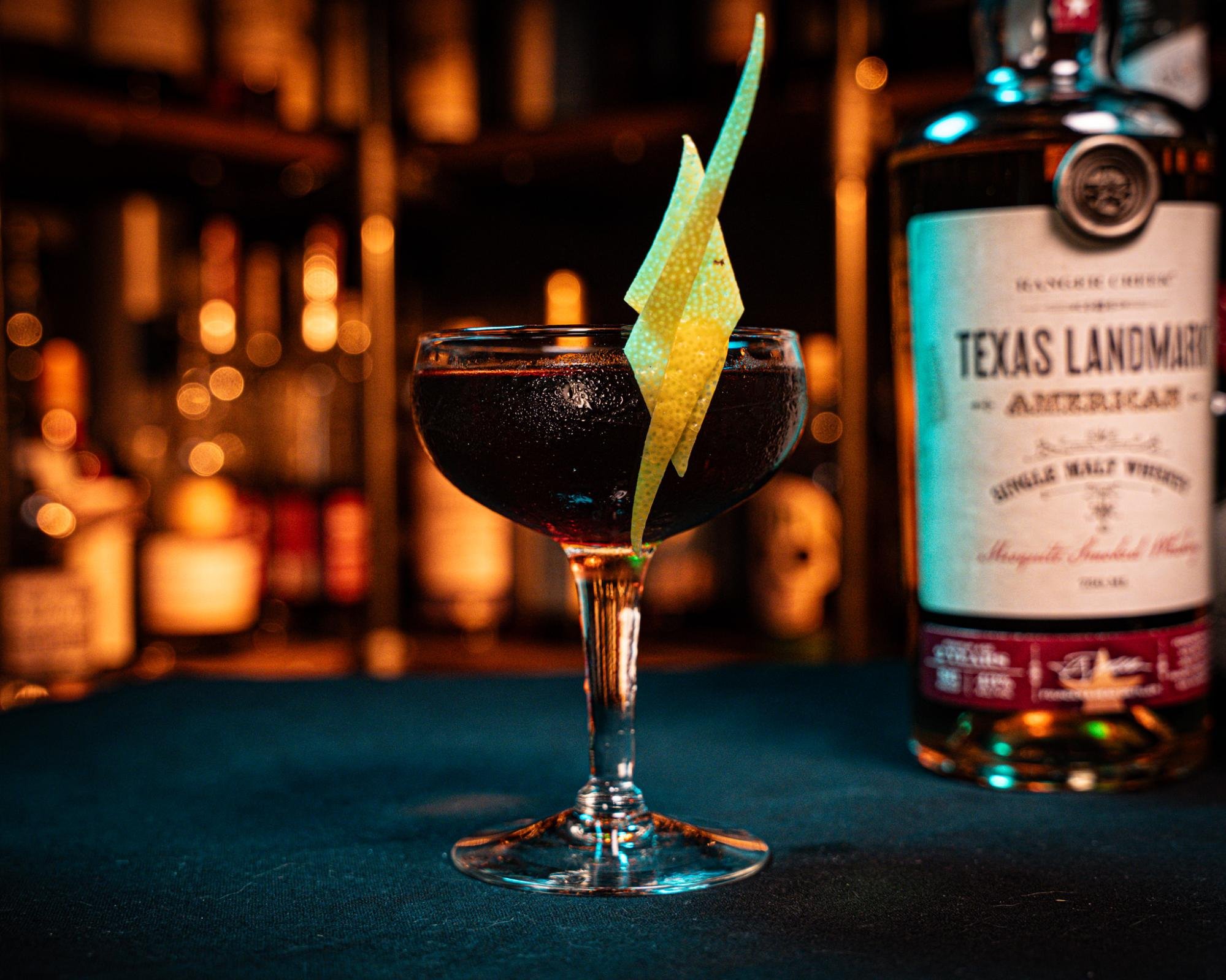The Sentimental Texan cocktail with a lemon twist beside a bottle of Texas Landmark single malt whiskey.