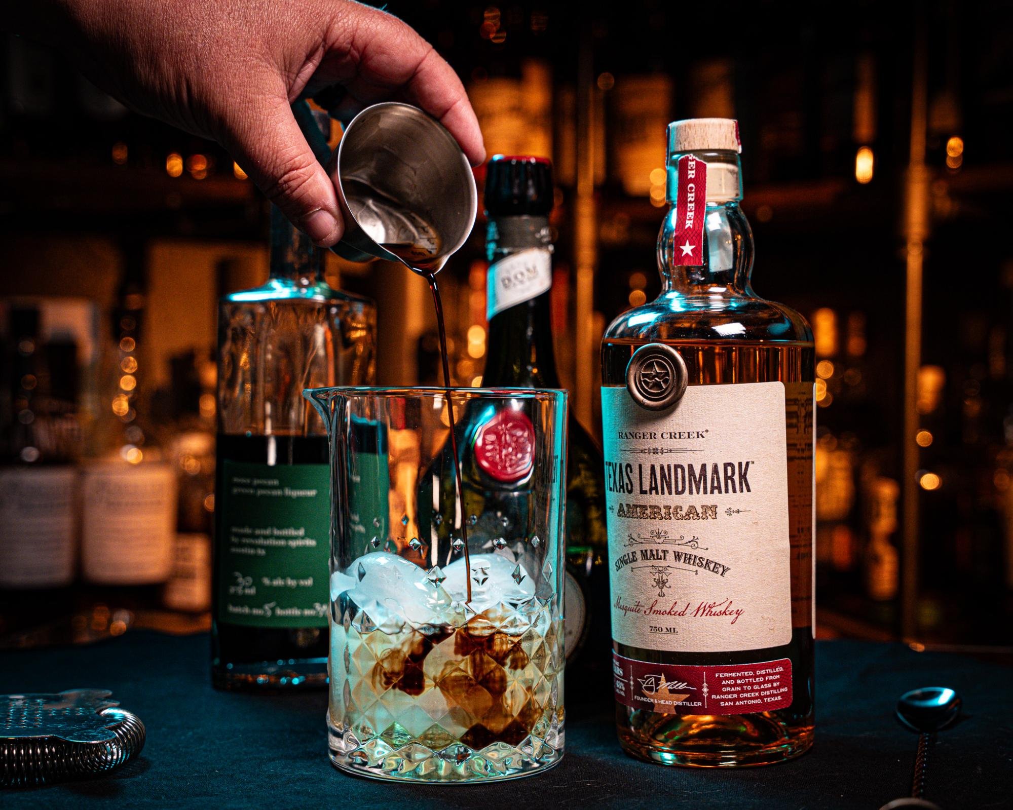 A measured ingredient is poured into a mixing glass beside the Sentimental Texan ingredient bottles.