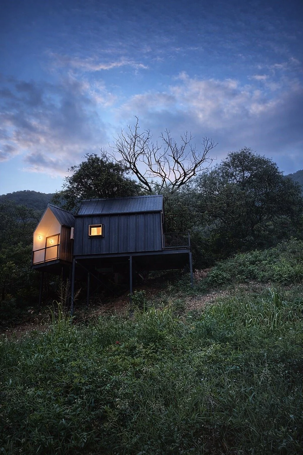 Casa de madera en un entorno natural, iluminada desde adentro, con árboles y una noche despejada con algunas nubes en el cielo.