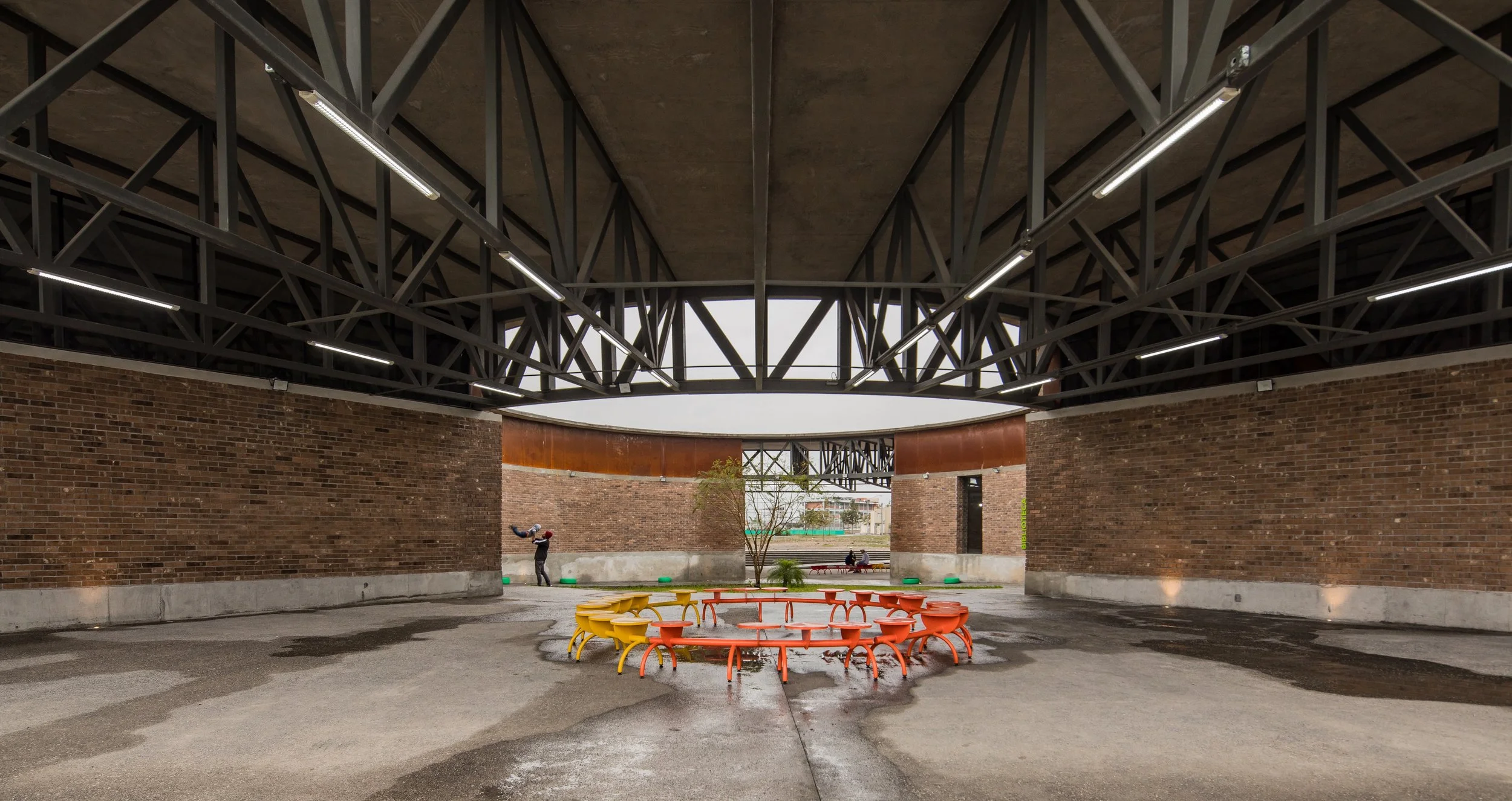 Ático con estructura metálica, paredes de ladrillo, mesas y bancos de colores, árbol en medio, personas en el fondo.