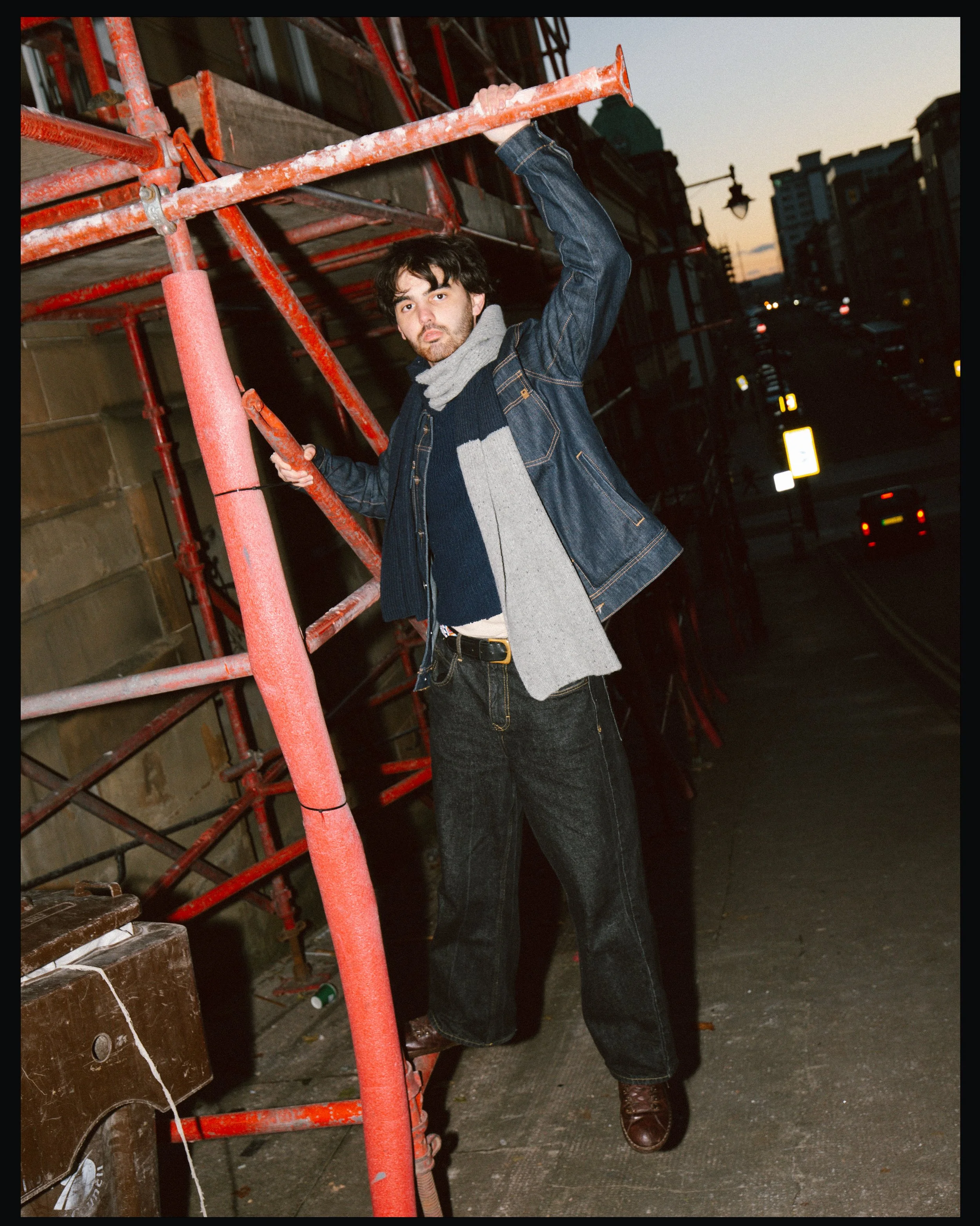 A young man standing on a construction scaffold on a city street during dusk, wearing a denim jacket, gray scarf, dark jeans, and brown boots.