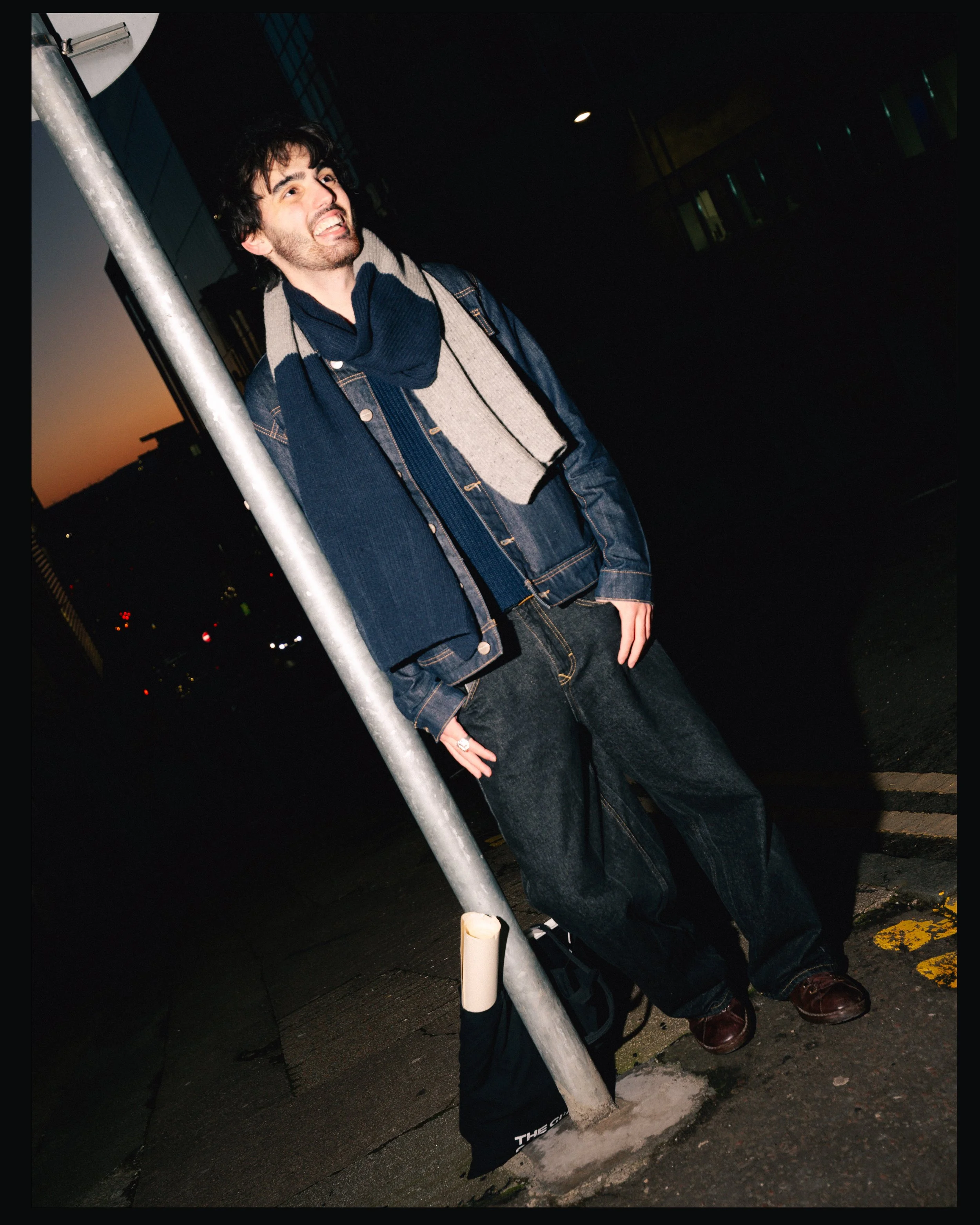 A young man standing next to a street pole during dusk, smiling and looking off into the distance. He is wearing a denim jacket, scarf, and jeans.