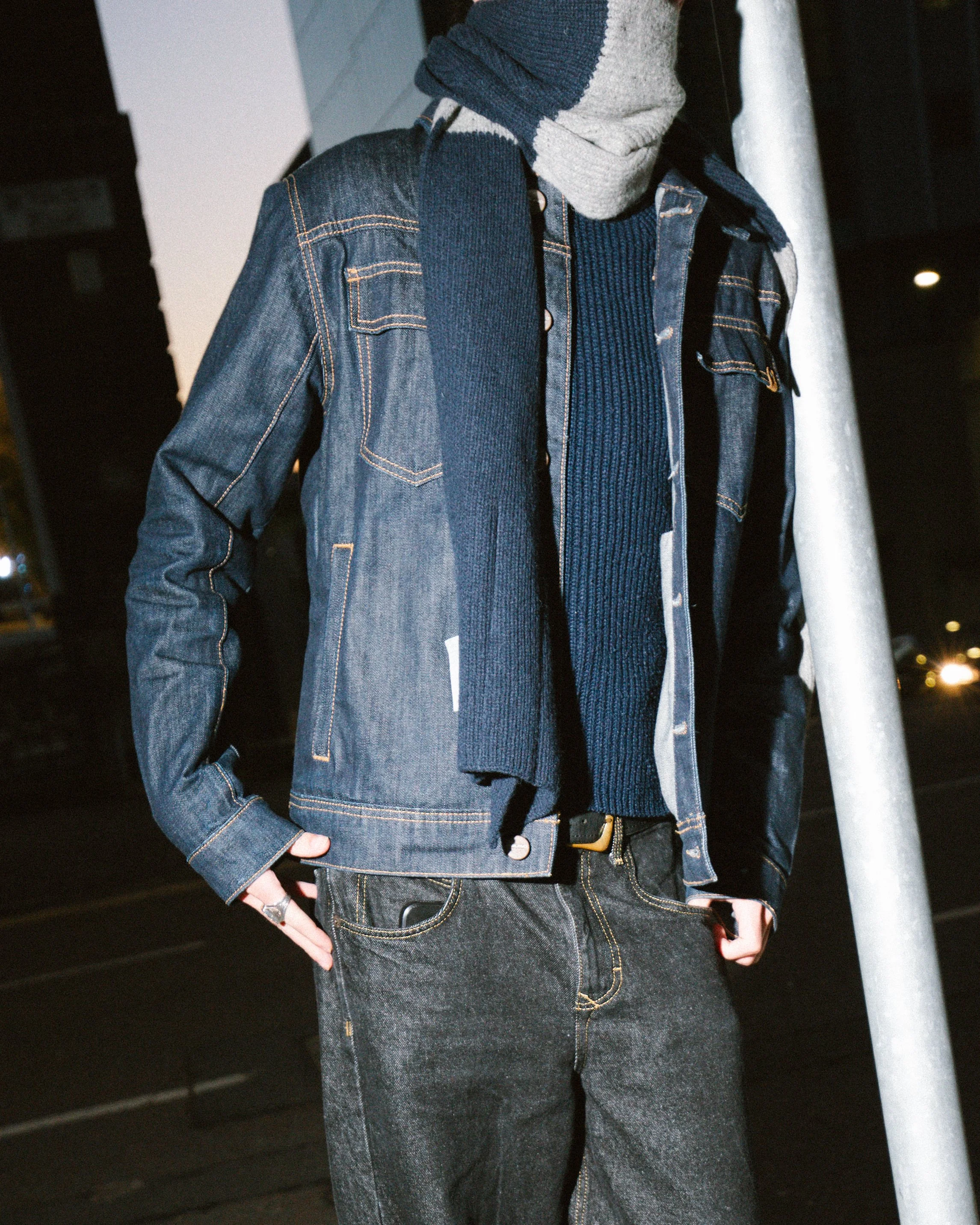 Close-up of a person dressed in dark denim jacket, dark jeans, and layered sweaters, standing outdoors at night.
