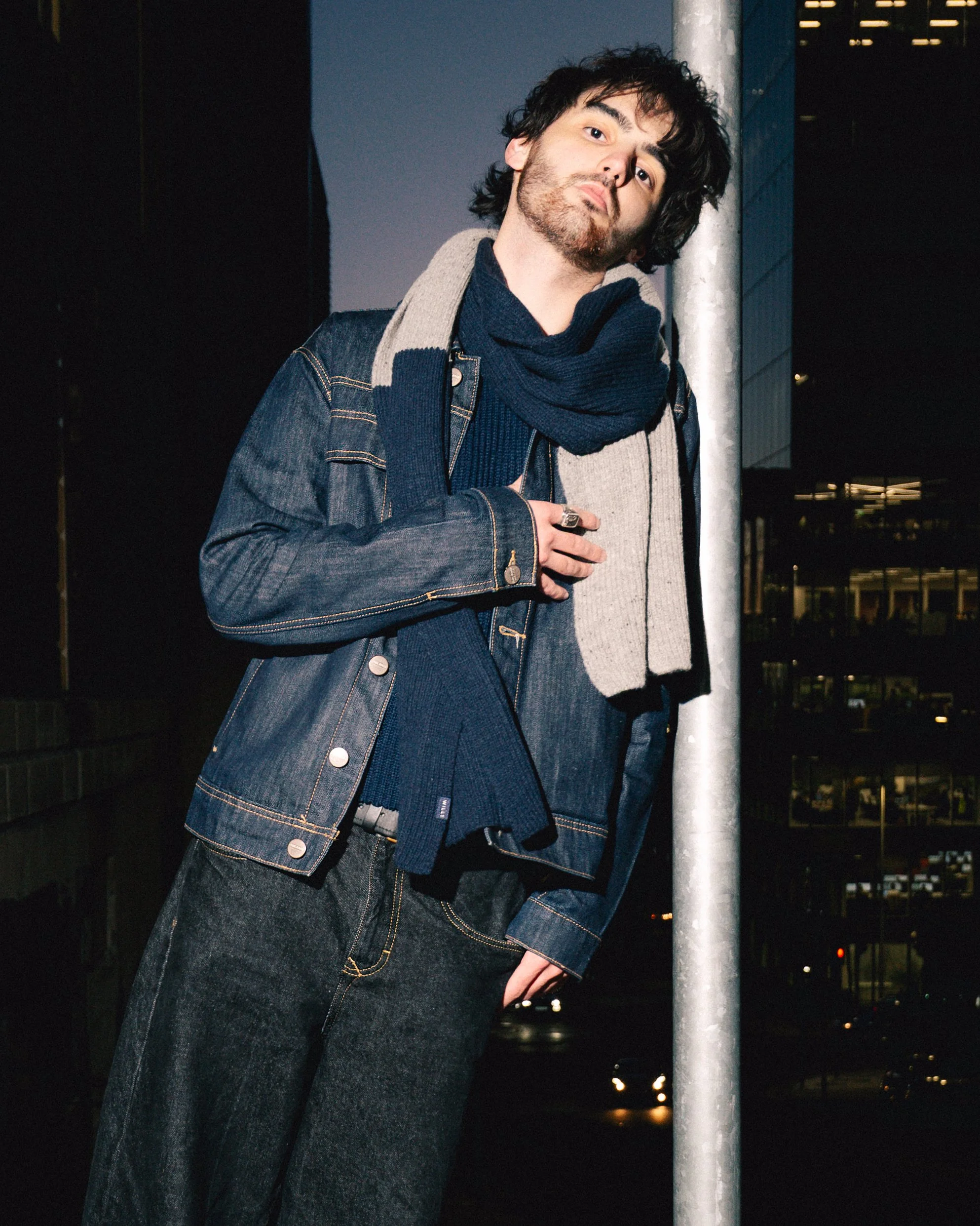 A young man with dark, curly hair and a beard standing outdoors at night near a metal pole, wearing a denim jacket, a navy sweater, a gray scarf, and dark jeans, with city lights in the background.