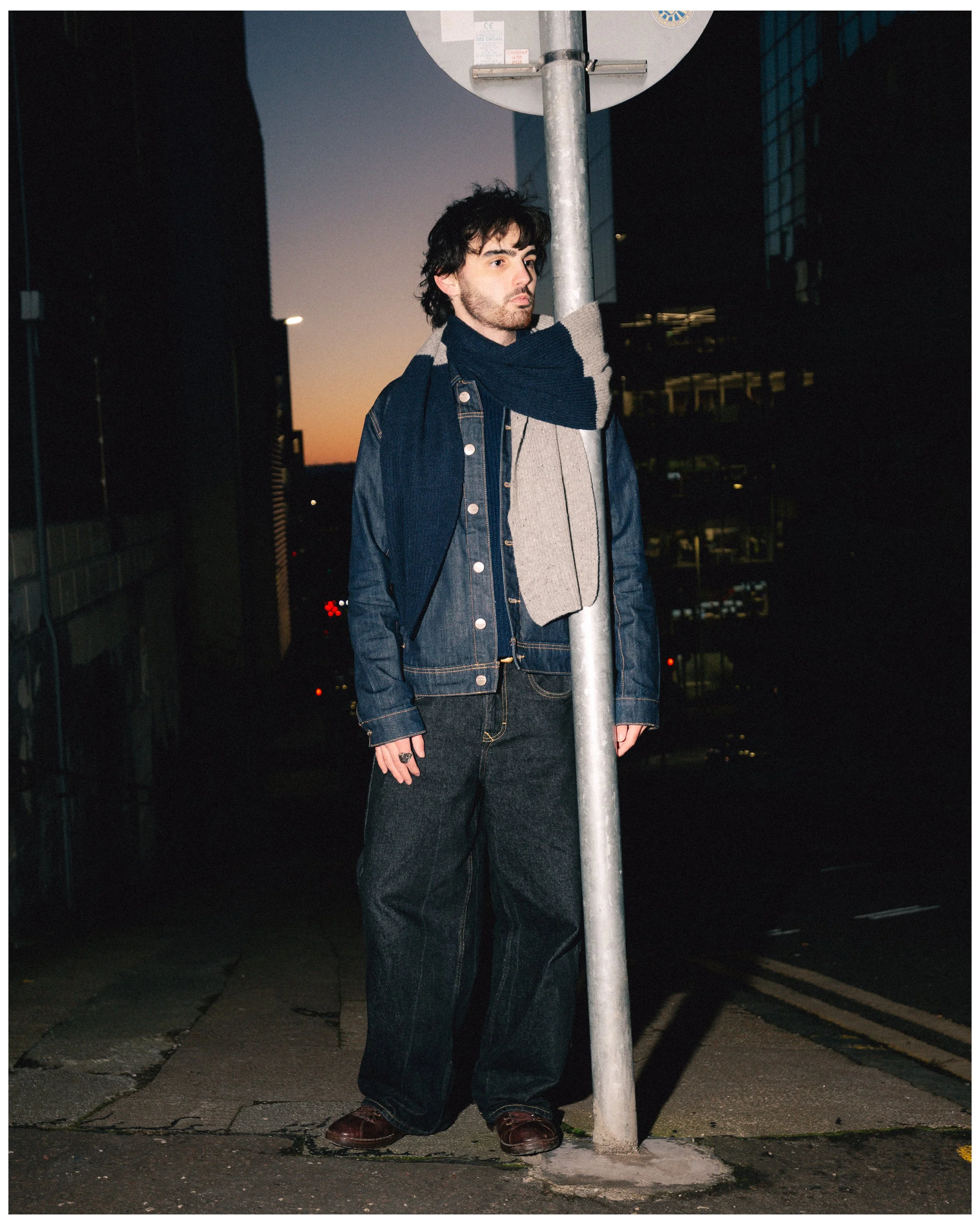 A man in dark jeans, a denim jacket, and a scarf stands next to a parking lot pole during dusk in an urban area.