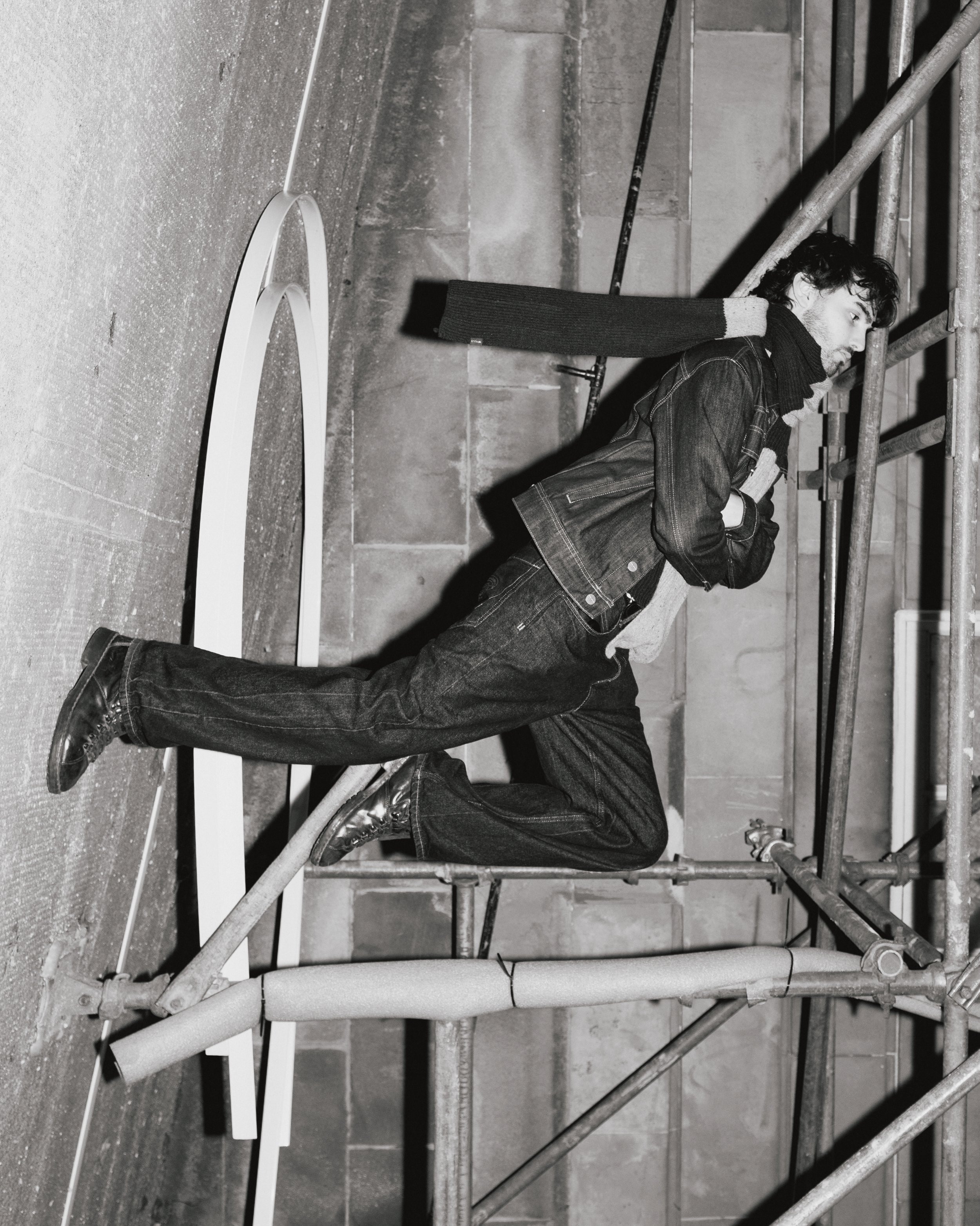 A man in dark clothing leaning on a scaffolding structure inside a room with concrete walls.