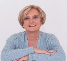Portrait of a smiling woman with blonde hair wearing a blue cardigan, posing with her arms crossed against a plain light background.