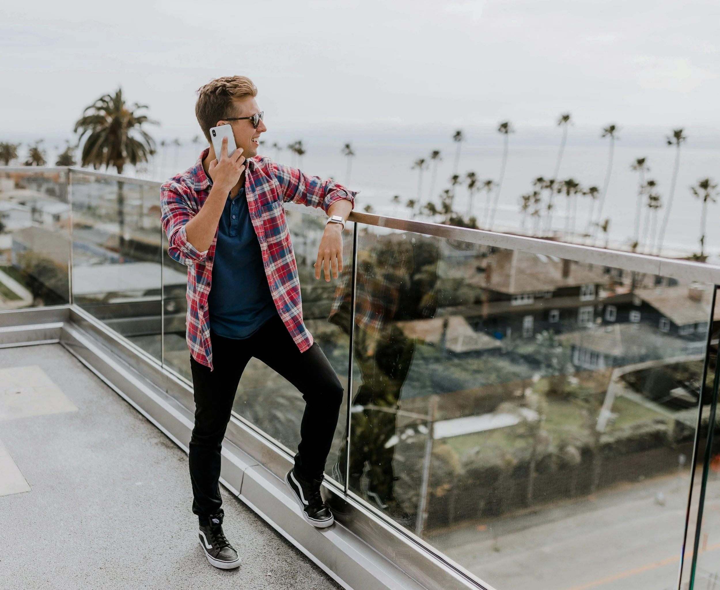 Young man in plaid shirt and sunglasses talking on cell phone on balcony with a view of palm trees and houses in the background.
