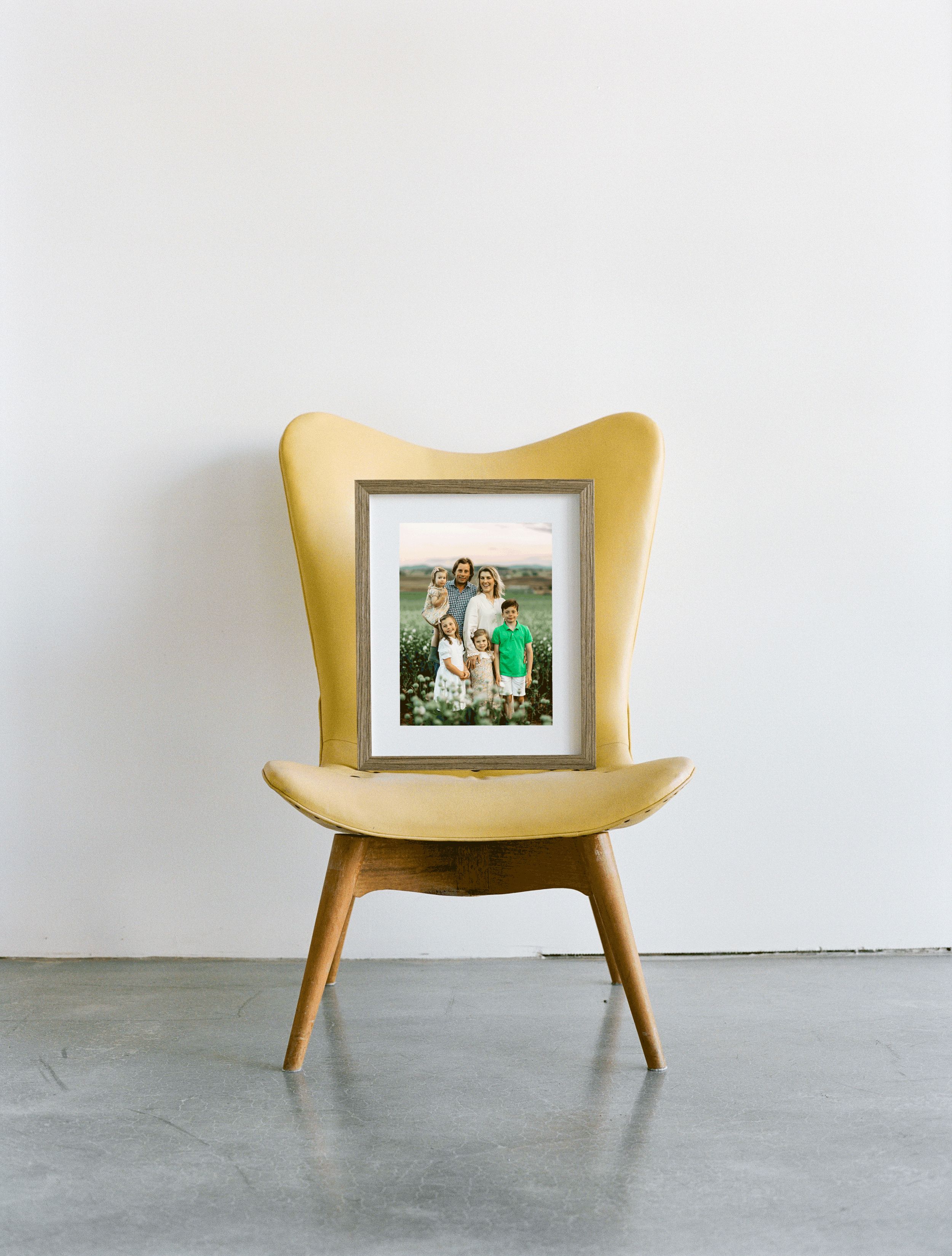 A modern yellow chair with a wooden frame placed on a gray floor, featuring a framed family photo resting on the seat against a white wall.