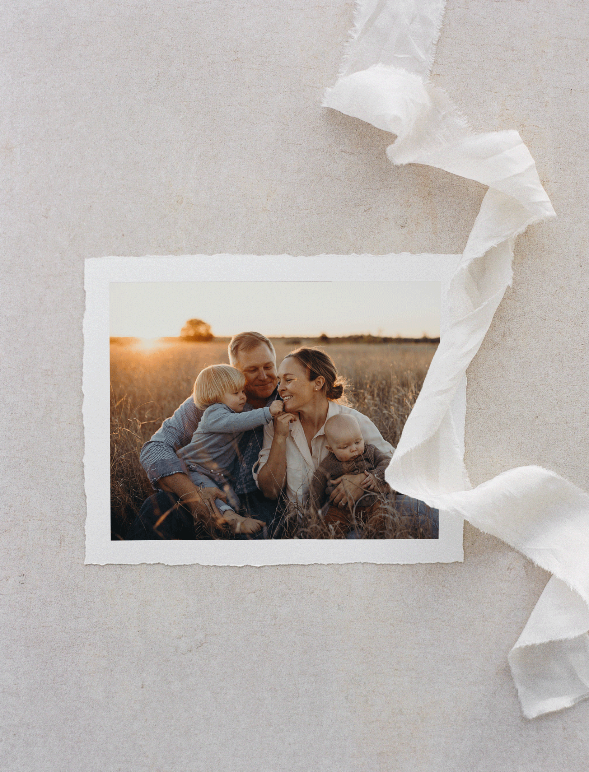 Family portrait with smiling parents and two young children in a field at sunset, surrounded by a decorative ribbon.