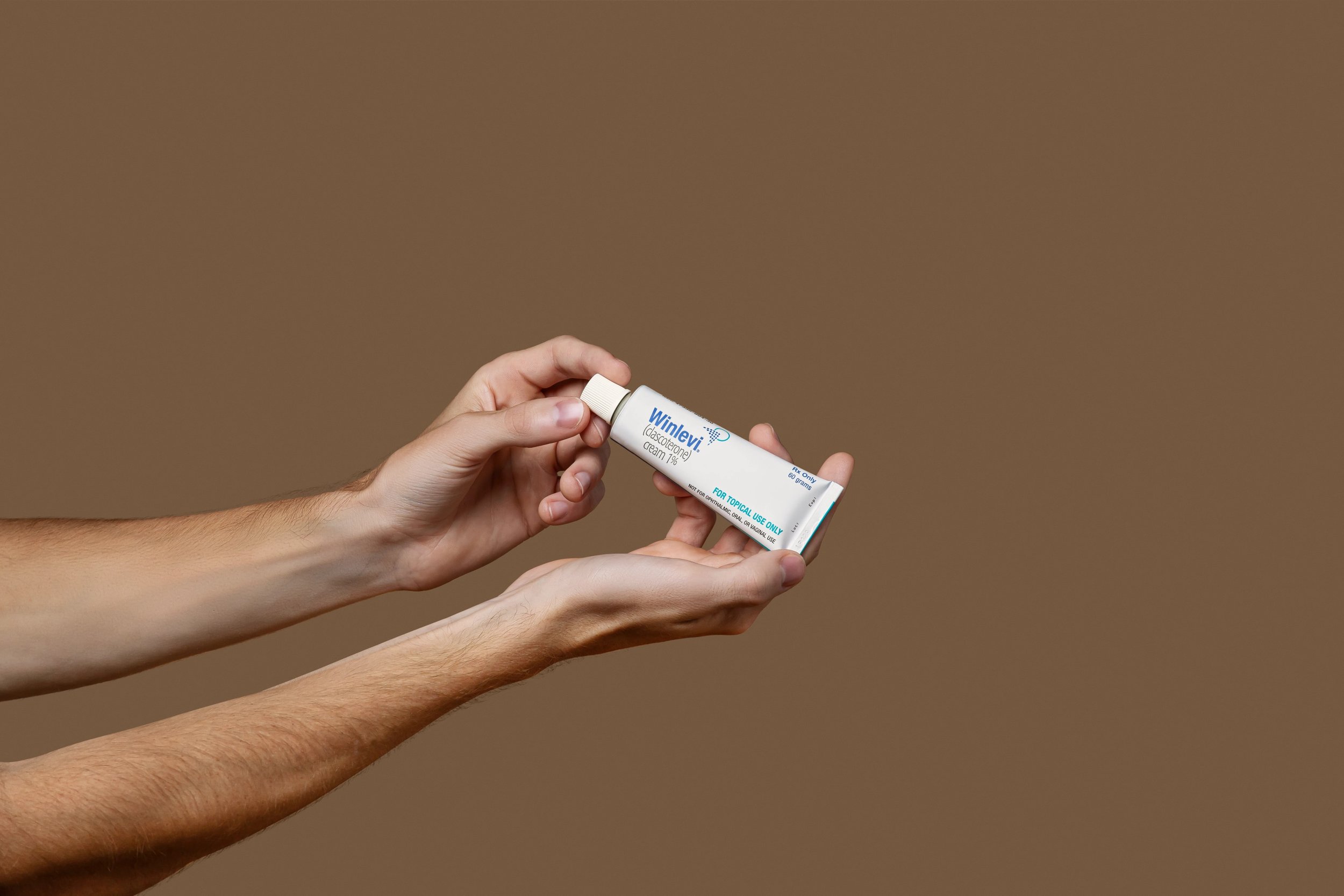 Hands holding a tube labeled Winlevi cream with clascoterone medication information on a brown background.