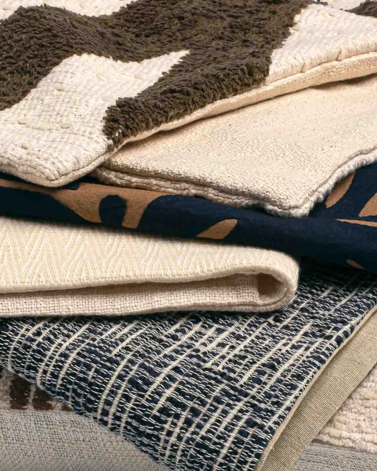Stack of folded fabrics with various textures and patterns, including a fuzzy brown and cream rug, a plain cream cloth, a navy fabric with beige patterns, and a black and white woven textile.