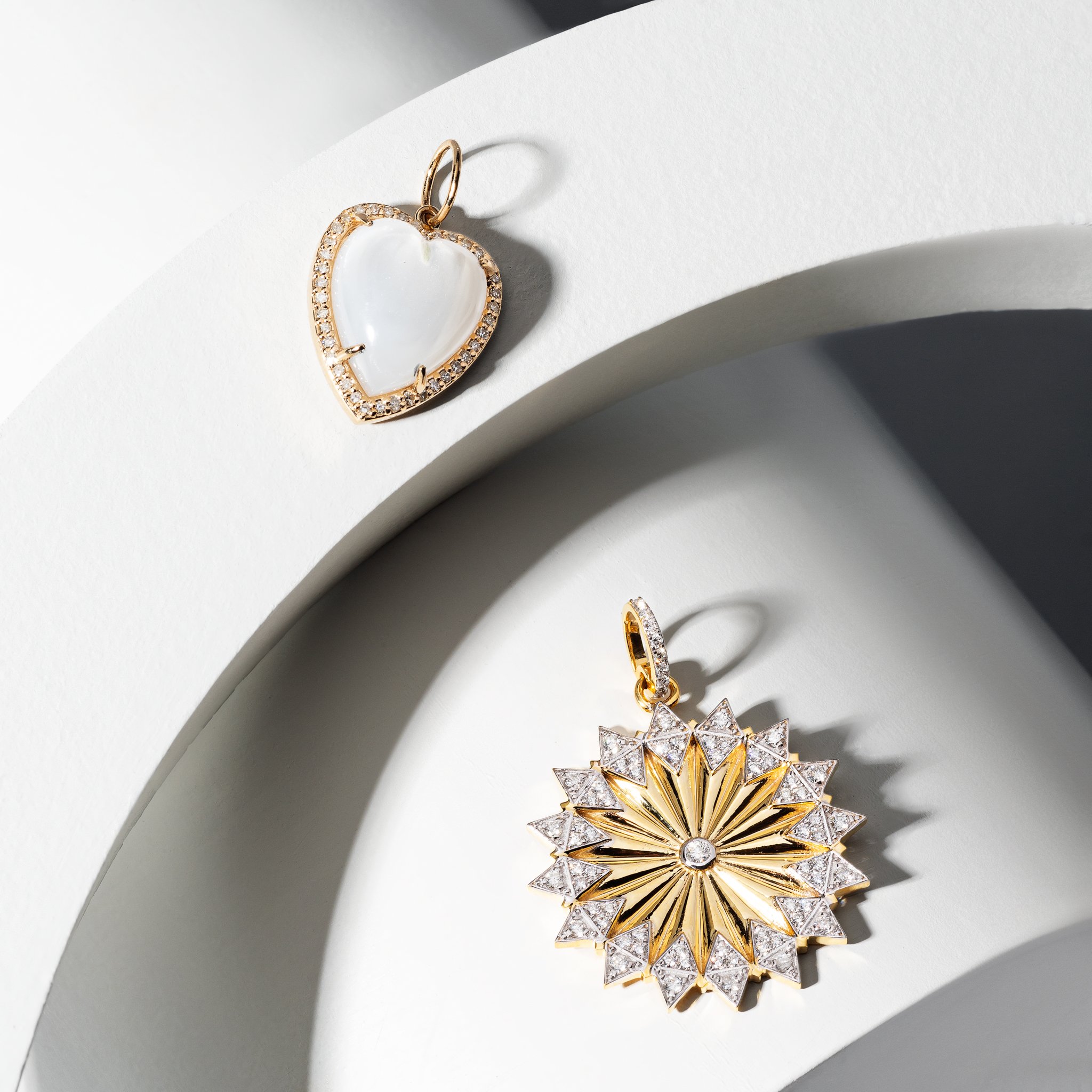 Two gold pendants with diamond accents on white surfaces; one is heart-shaped with a mother-of-pearl center, the other is a sunburst design with a star pattern.