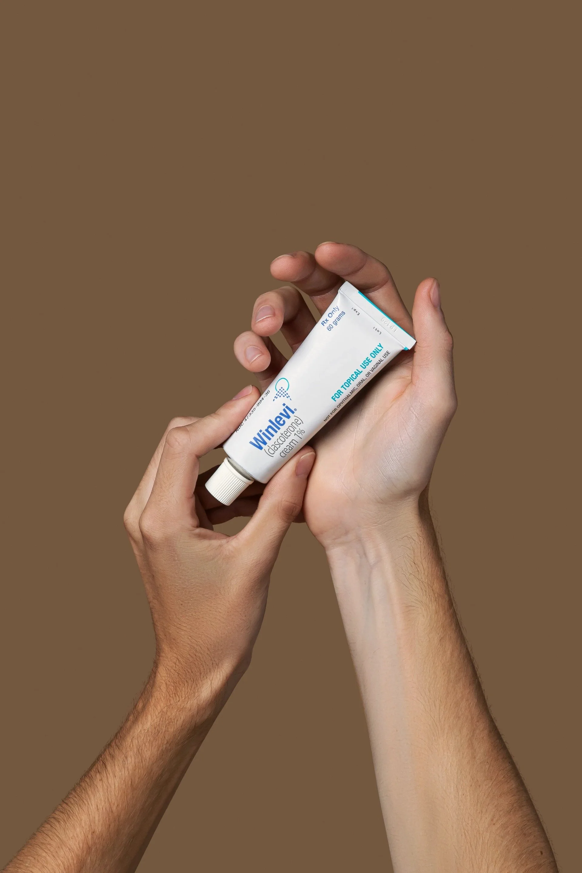 Hands holding a tube of Winlevi topical cream against a brown background.