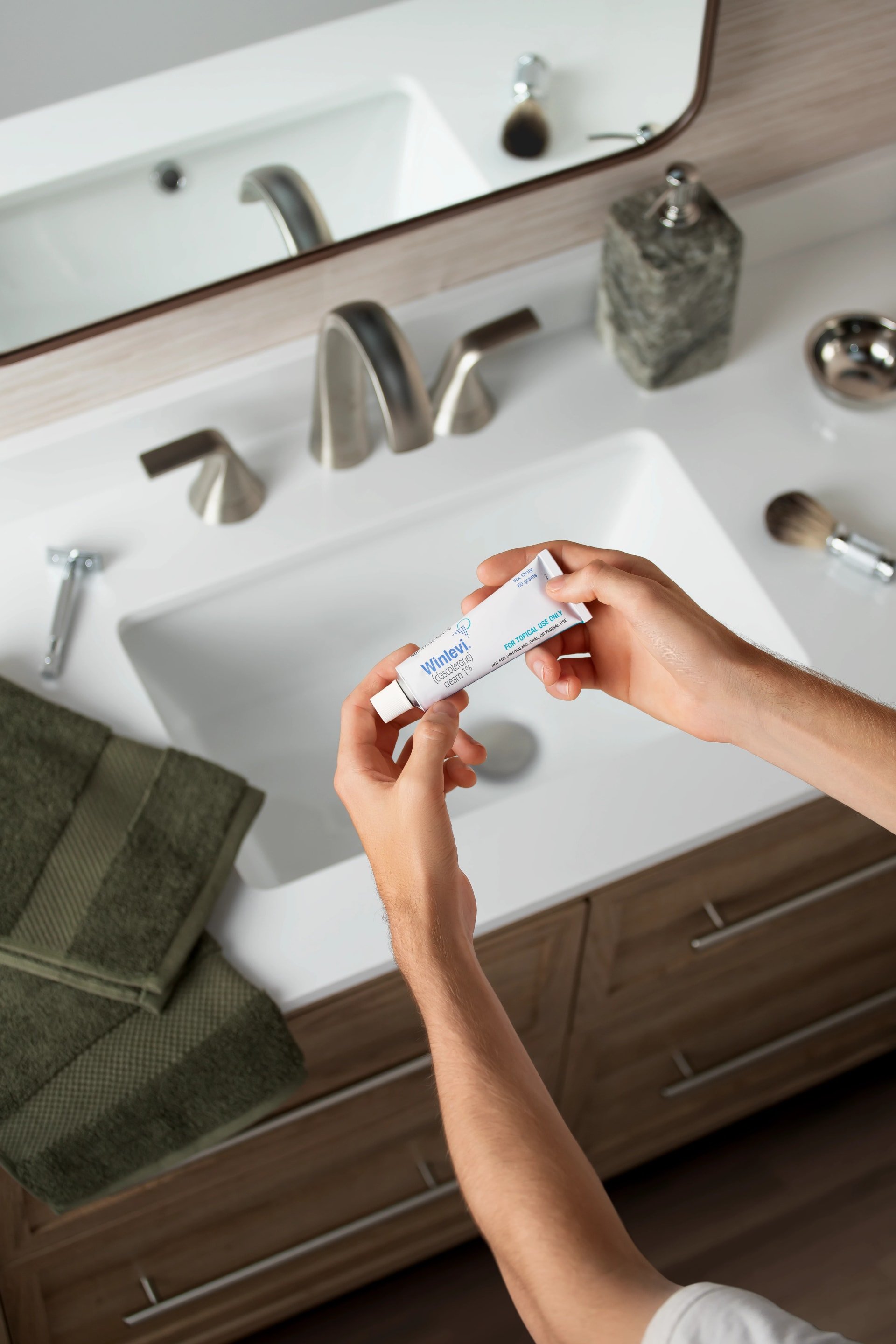Hands holding a tube of Winlevi cream over a bathroom sink, with a green towel and grooming items nearby.