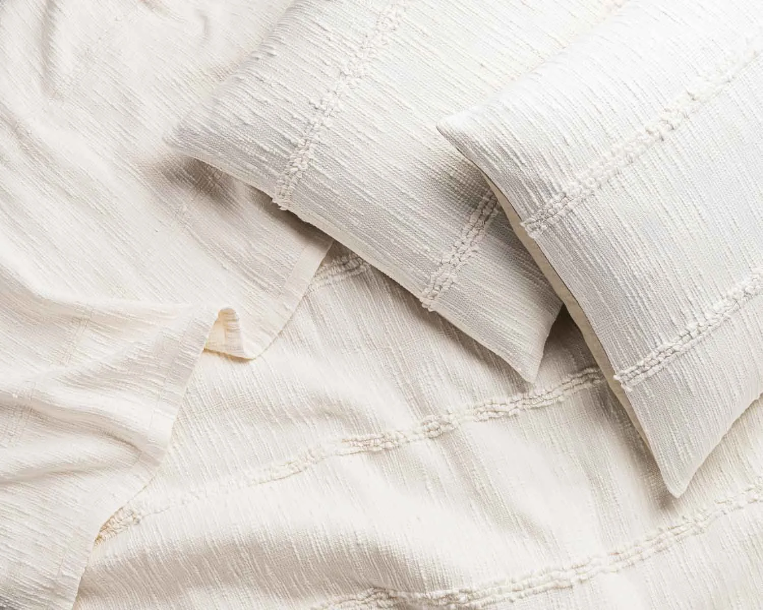 Close-up of cream-colored fabric with textured lines and embroidered details, possibly a bedsheet and pillowcase.