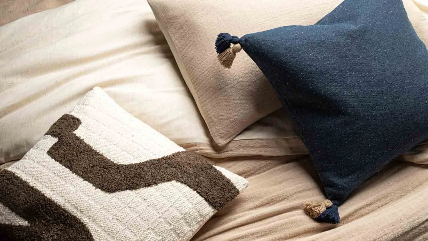 Three decorative pillows arranged on a bed with beige and light brown bedding, including a dark blue pillow with tassels, a beige pillow, and a textured pillow with a brown and white geometric pattern.