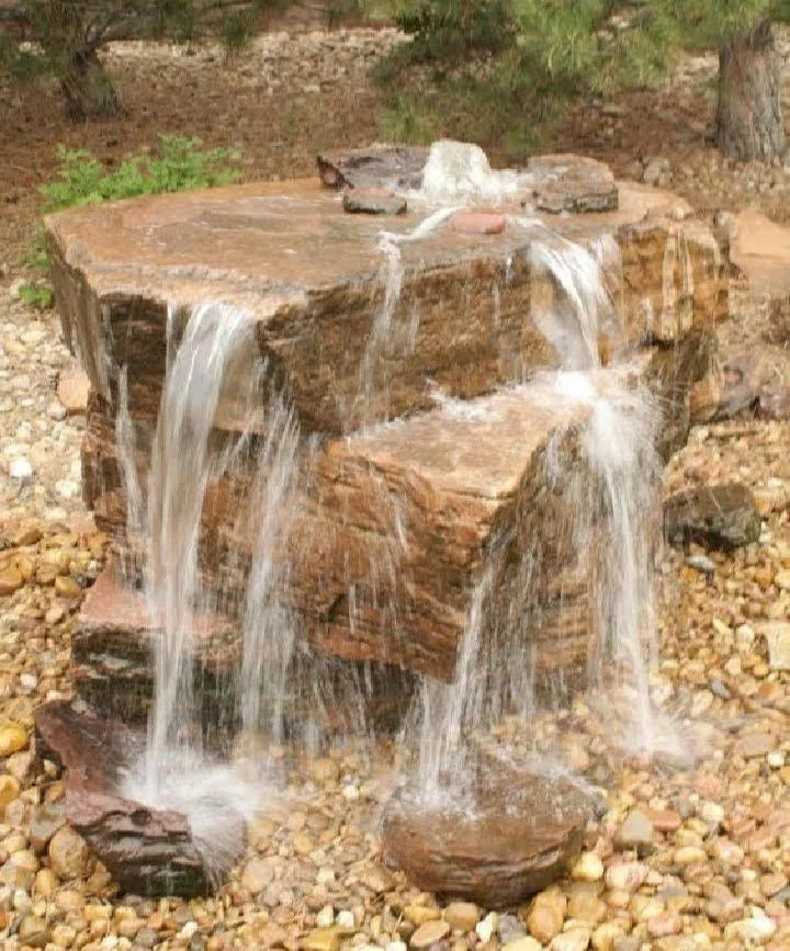Water flows over stacked large flat rocks, creating a small waterfall on a gravel surface in an outdoor setting.
