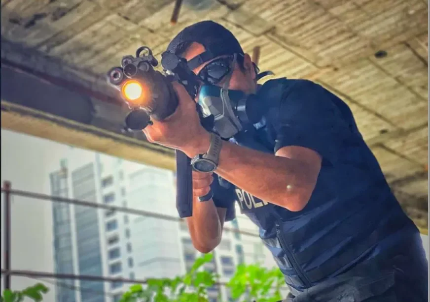 A police officer in tactical gear, including a face mask and goggles, aiming a rifle with an attached flashlight at an unseen target, indoors or under a bridge.