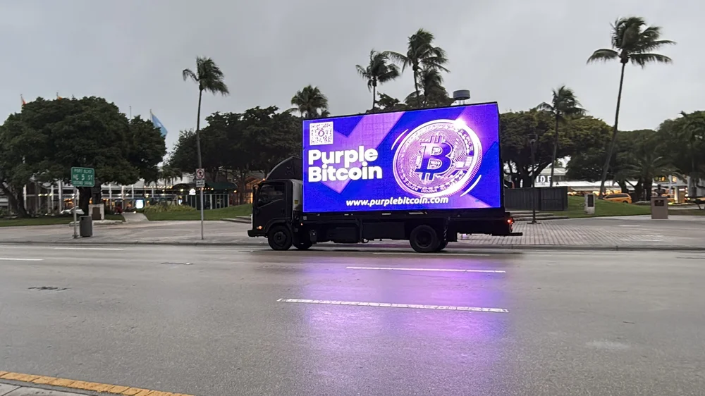 Mobile LED billboard truck displaying a Purple Bitcoin advertisement near palm trees