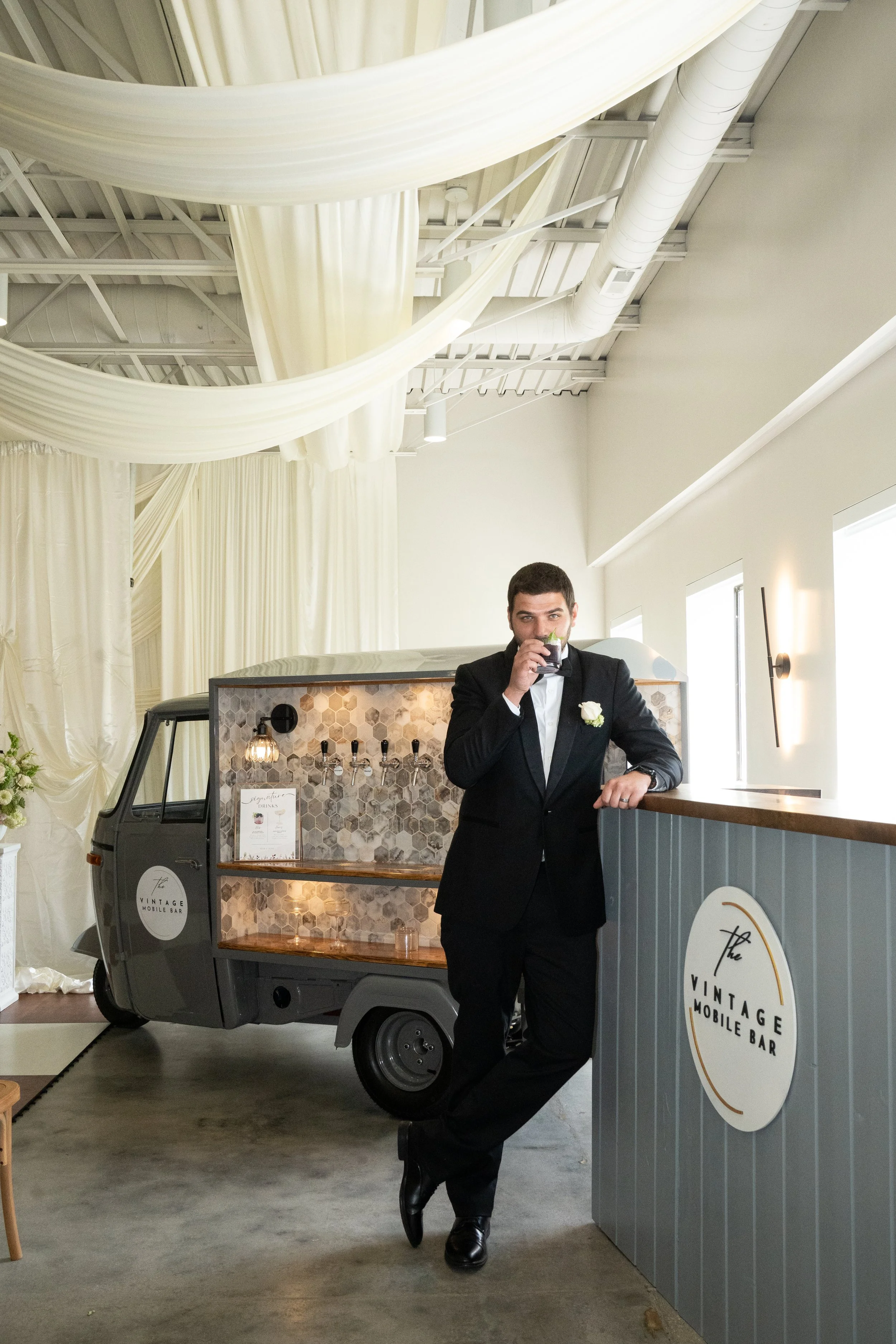 A man dressed in a black tuxedo with a white shirt and black bow tie, standing next to a mobile bar shaped like a vintage truck. He is holding a drink and leaning against a counter. The mobile bar has a sign that reads "The Vintage Mobile Bar." There is a decor setup with cream-colored drapes and large windows in the background.