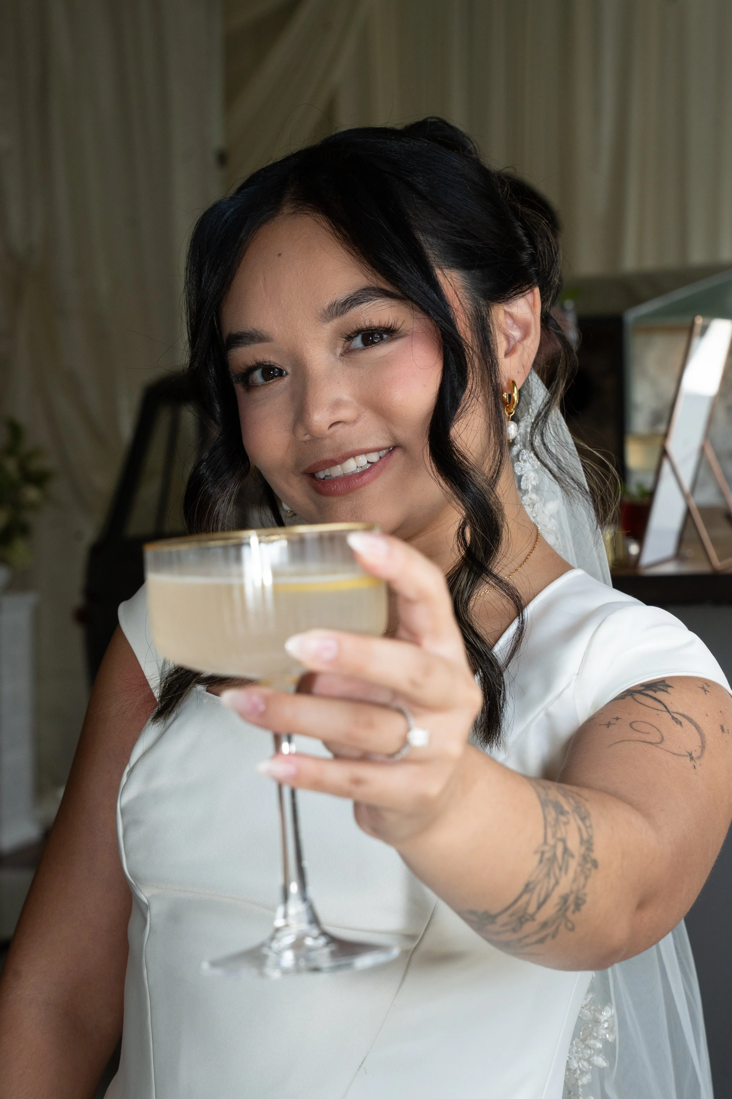 Close-up of a smiling woman with dark hair and tattoos, holding a cocktail glass in front of her, wearing a white dress and earrings, in an indoor setting.
