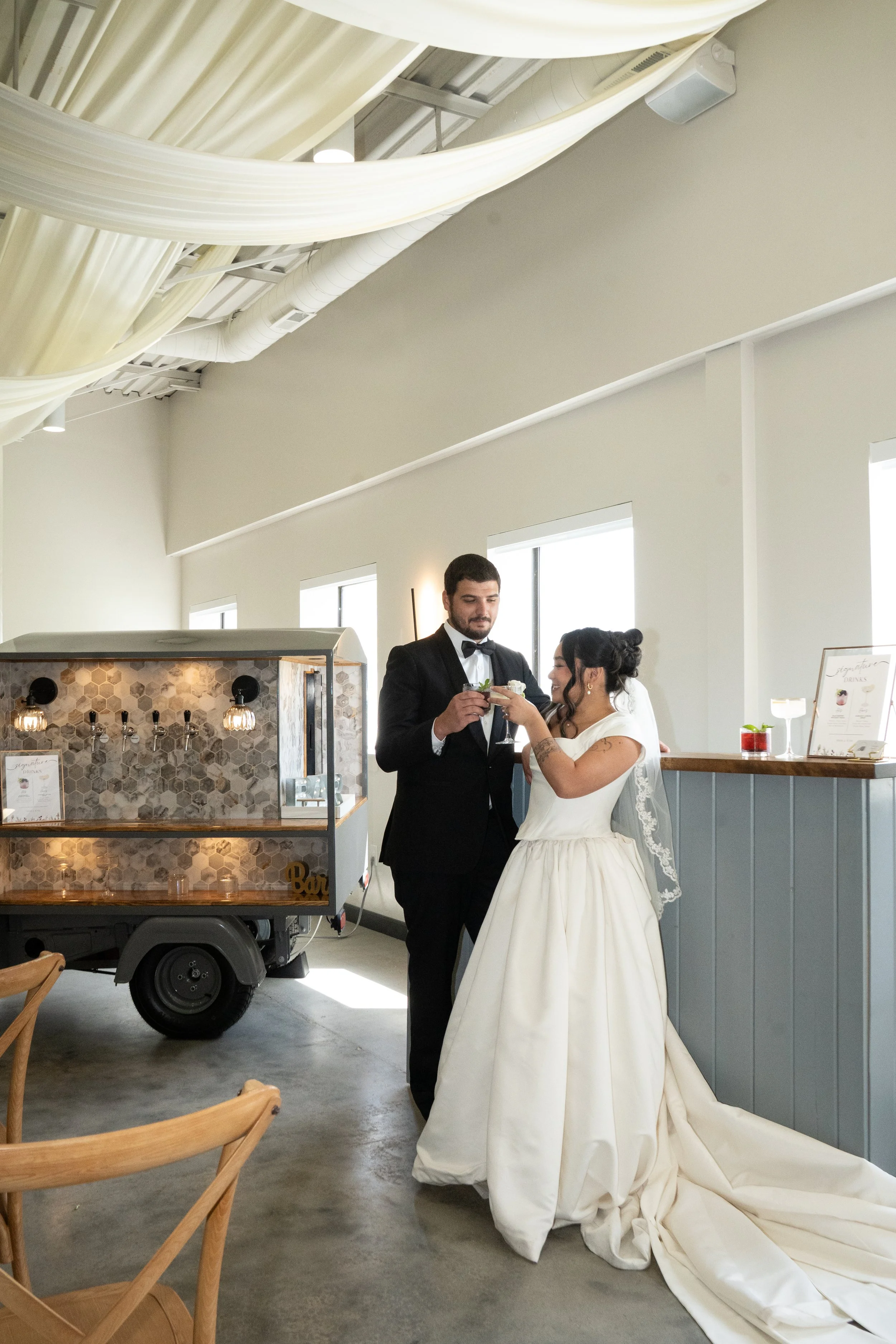 A bride and groom in wedding attire sharing a toast inside a modern venue. The bride is in a white gown and the groom in a black tuxedo. There's a mobile bar with a honeycomb tile design and a small menu on a stand nearby. The ceiling has exposed ductwork and fabric drapes, with large windows letting in natural light.