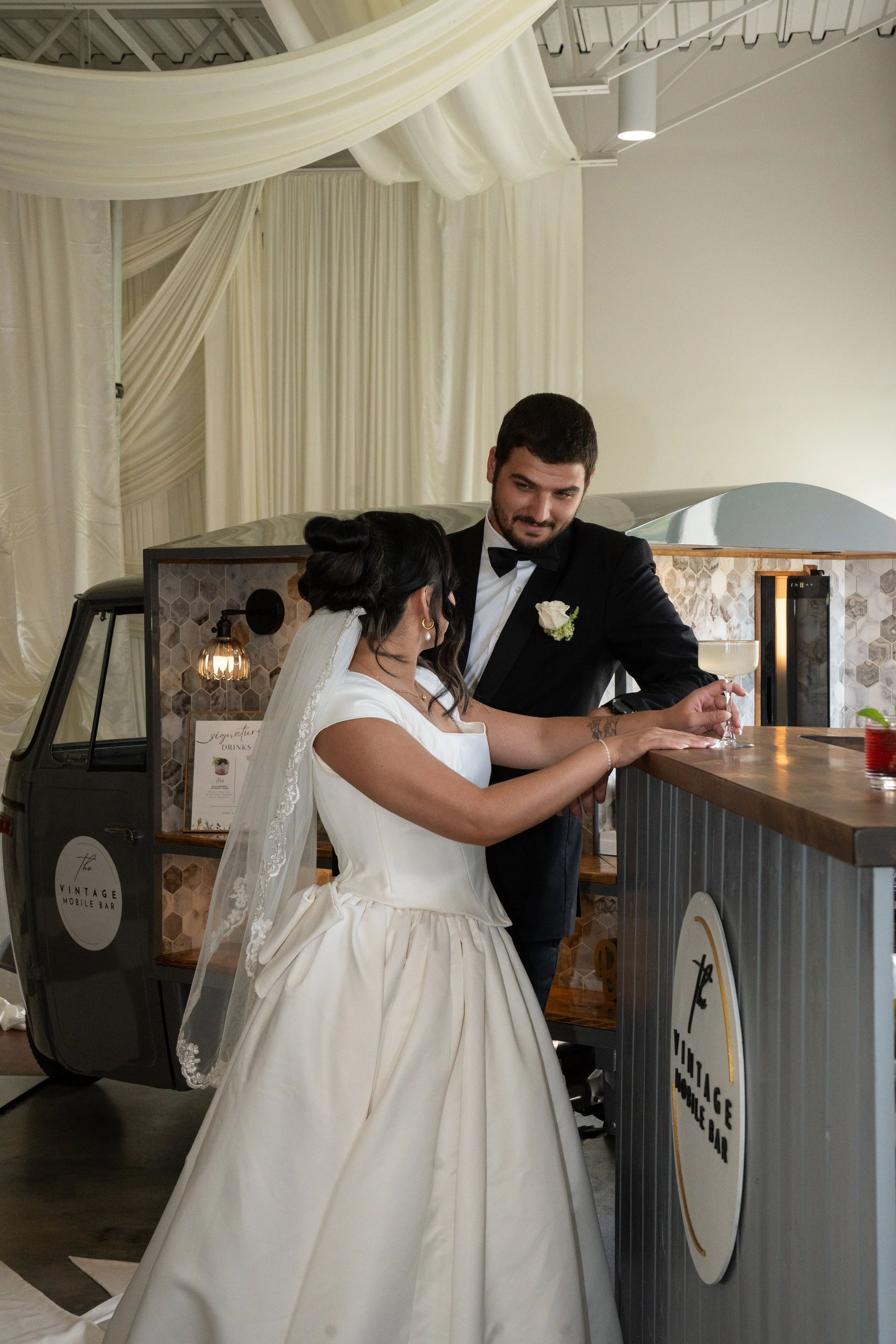 A bride in a white wedding dress and veil standing at a bar, talking to a groom in a black tuxedo with a white boutonnière, holding a cocktail glass in a decorated event space with draped curtains overhead.