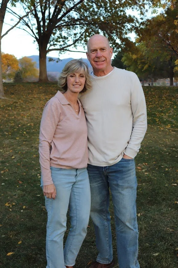 A middle-aged woman and a middle-aged man standing outdoors in a park with autumn foliage, smiling at the camera.
