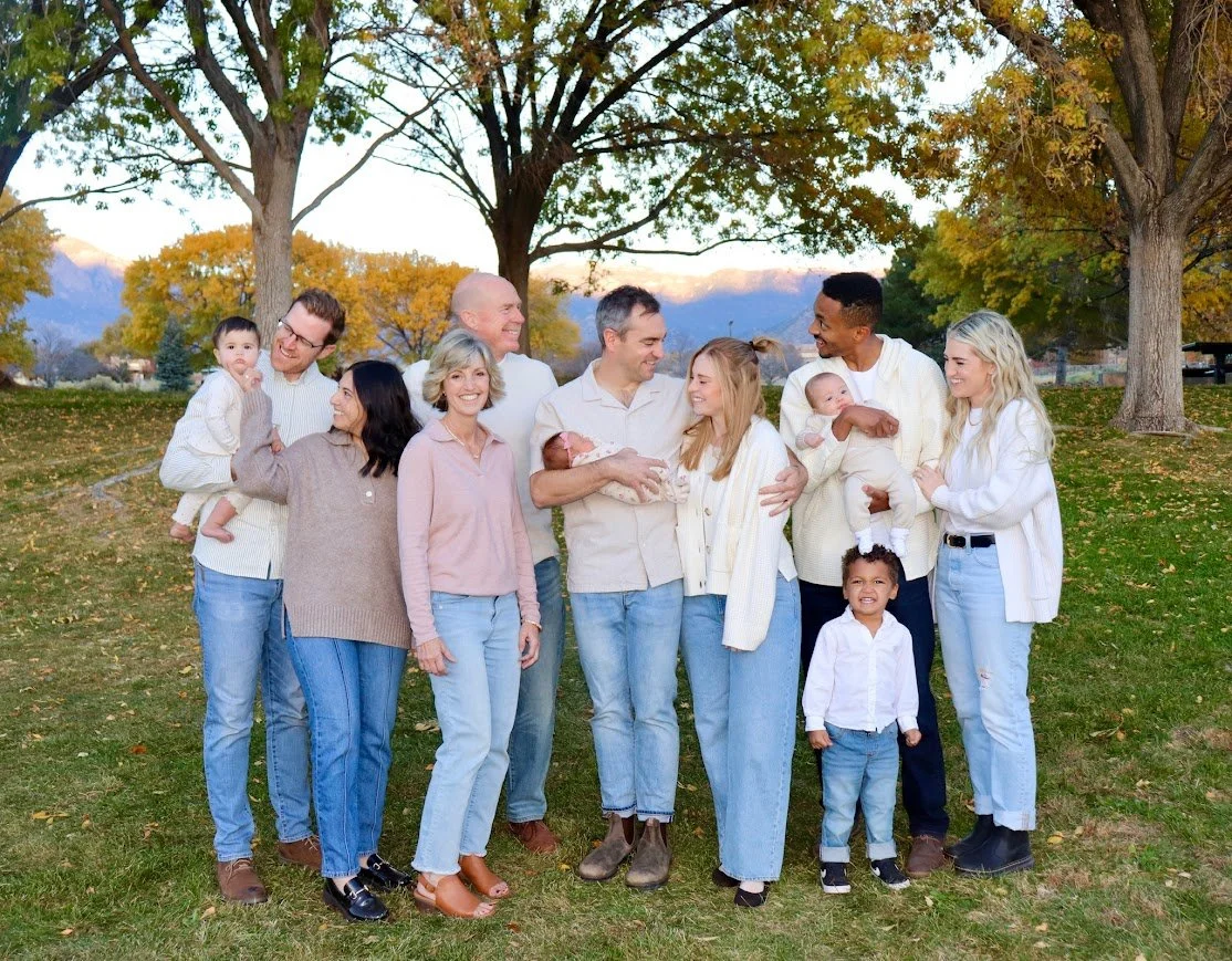 Family of thirteen members standing outdoors in a park with autumn foliage, smiling and holding babies and young children.