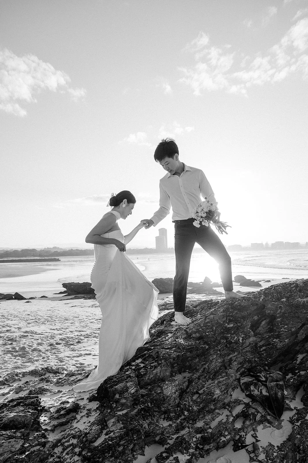 A couple, dressed in wedding attire, stands on rocks on Currumbin beach at sunset. Photo taken by Gold Coast Engagement photographer Alix Dubois.