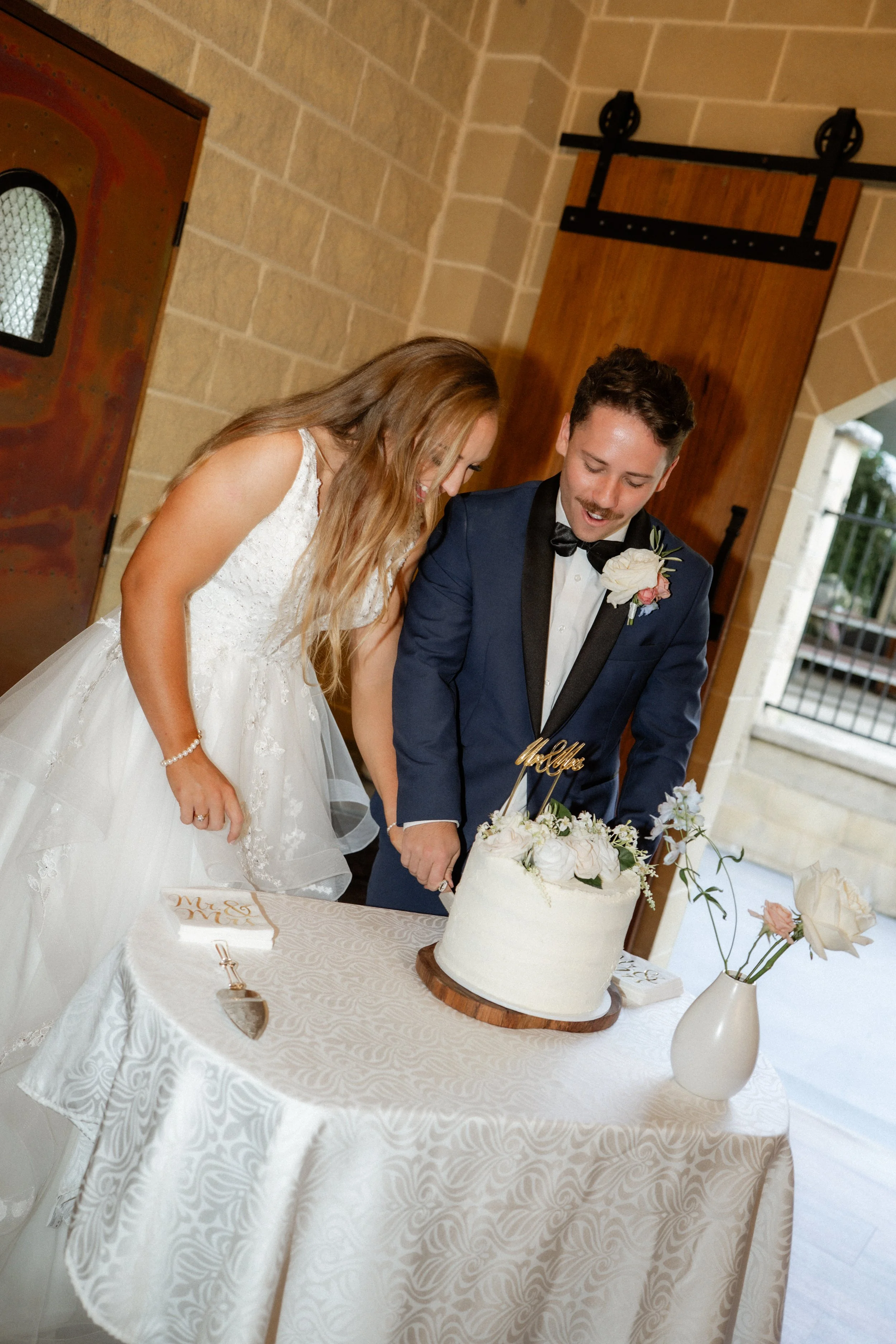 Bride and groom cutting wedding cake at reception, dressed in wedding attire, indoors with beige brick walls and a wooden sliding barn door.