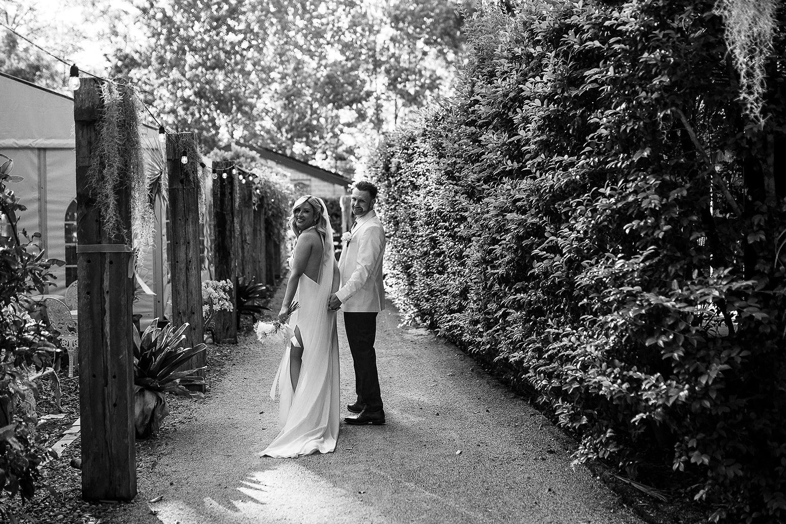 Black and white photo of a bride and groom holding hands and smiling, walking down a garden path surrounded by hedges and trees, with sunlight filtering through the leaves.