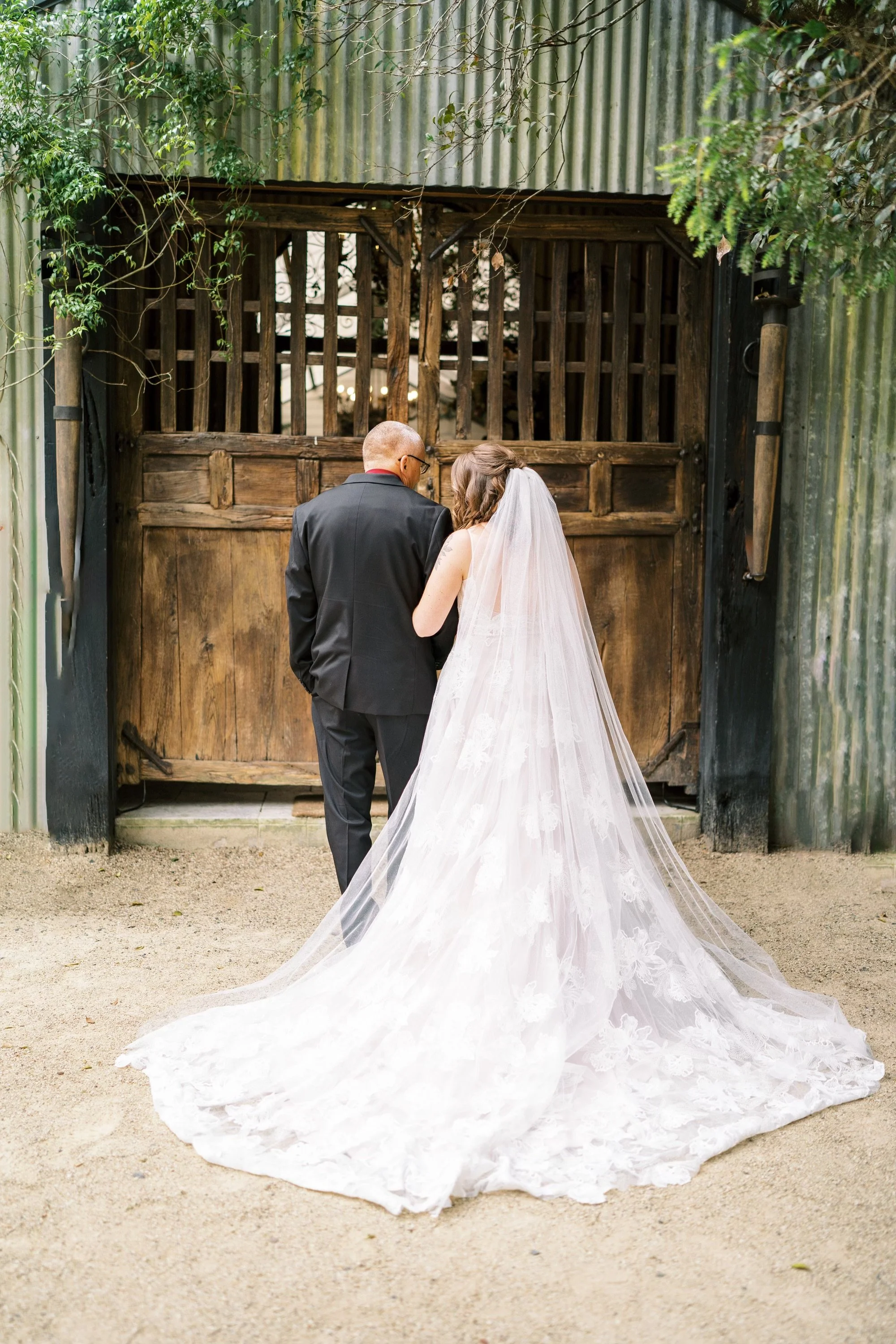 A bride in a white wedding dress with a long train and veil standing beside a man in a tuxedo, facing a large wooden gate in an outdoor setting surrounded by greenery.