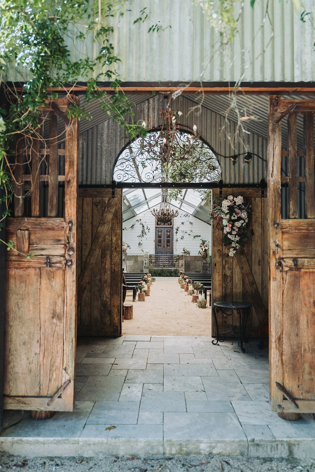 Rustic barn doors open to a decorated aisle with wood slices as seats, leading to a floral arch in a wedding venue with chandeliers and greenery.