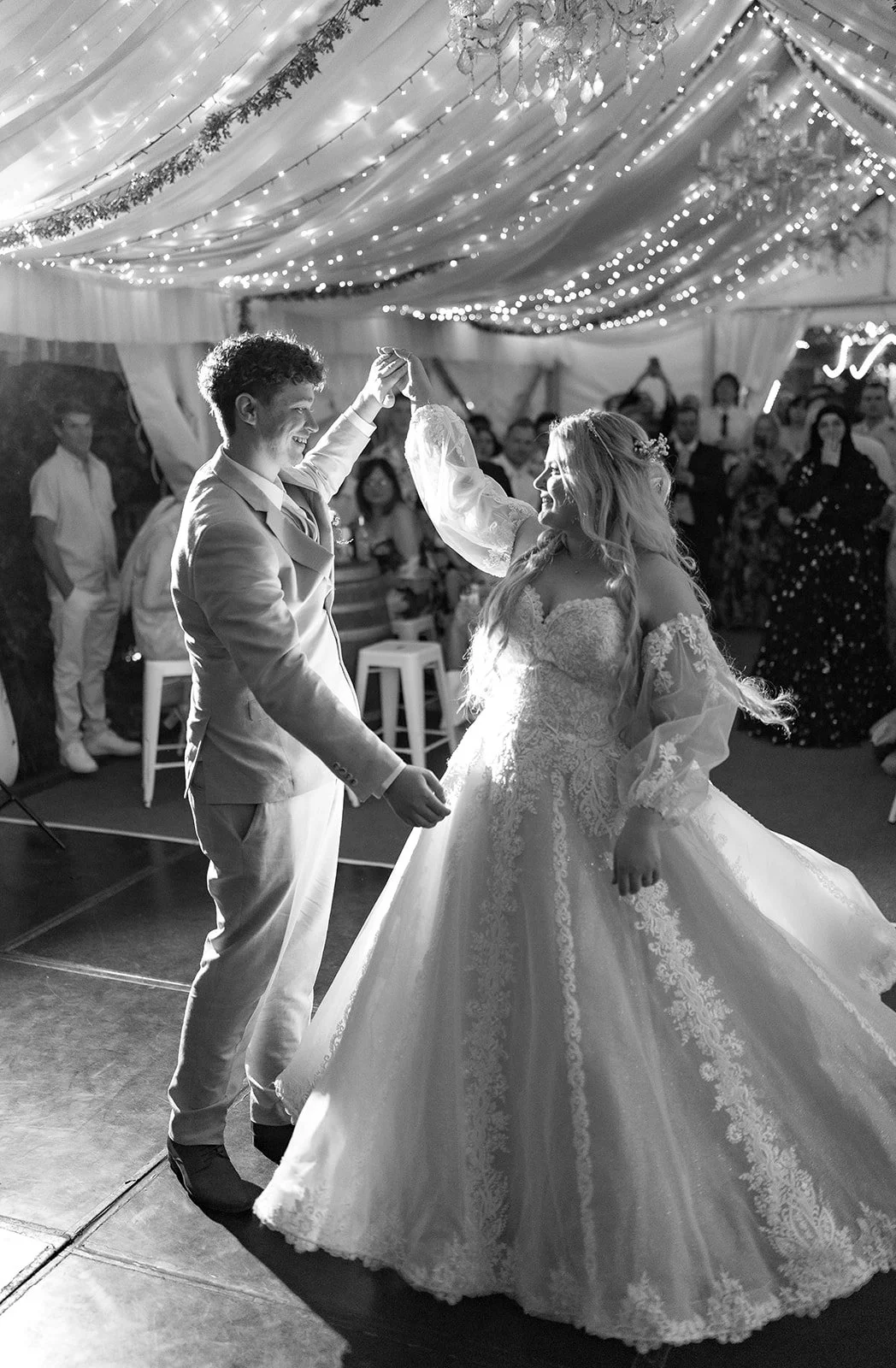 Black and white photo of a bride and groom dancing at their wedding reception, surrounded by guests and decorated with string lights and chandeliers.