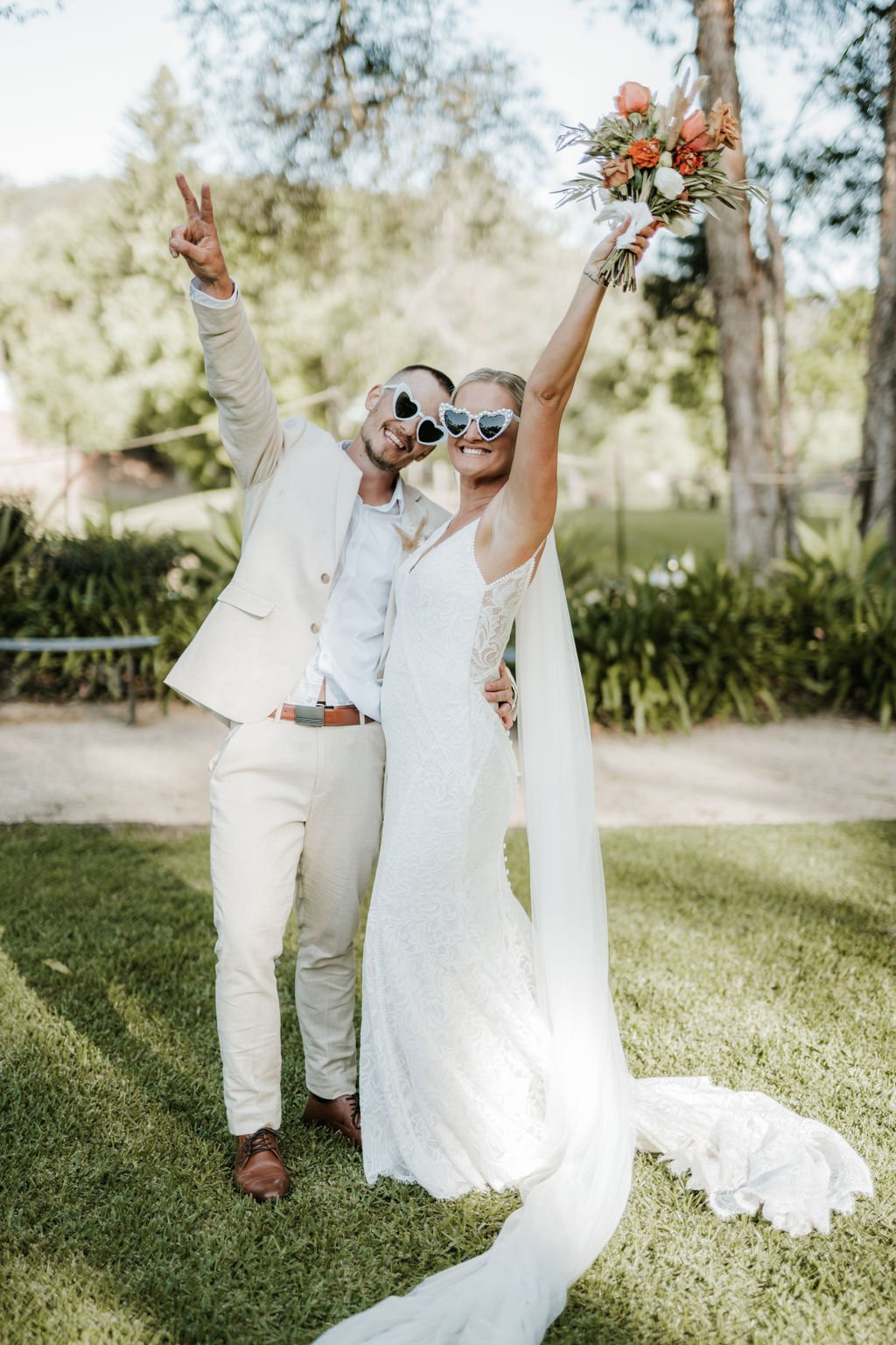 A joyful bride and groom celebrating outdoors, wearing sunglasses and holding a bouquet, with the bride raising her arm in excitement.