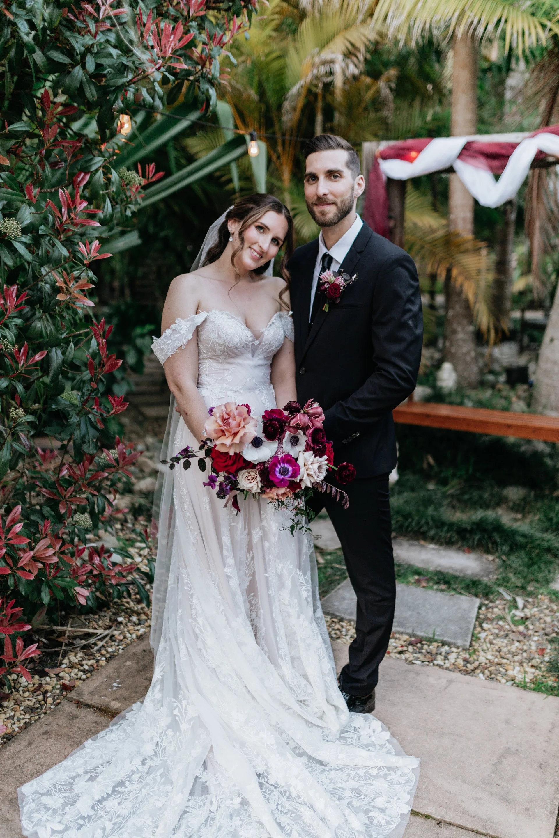 A bride and groom standing in a lush outdoor garden, with the bride holding a colorful bouquet of pink, purple, and white flowers. The bride wears a white off-shoulder lace wedding dress, and the groom is in a black suit with a boutonniere. They are smiling and posing for a wedding photo.