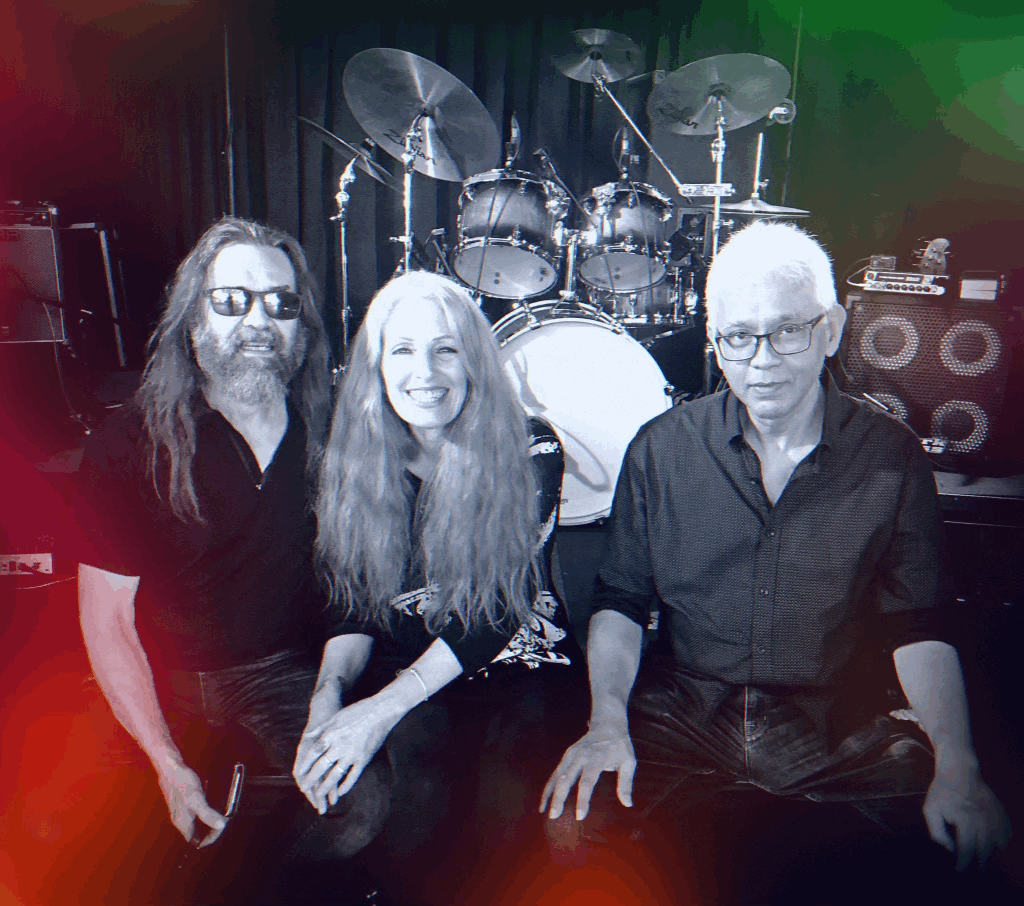 Three musicians sitting in front of drums and electrical equipment on stage, with colorful lighting effects.