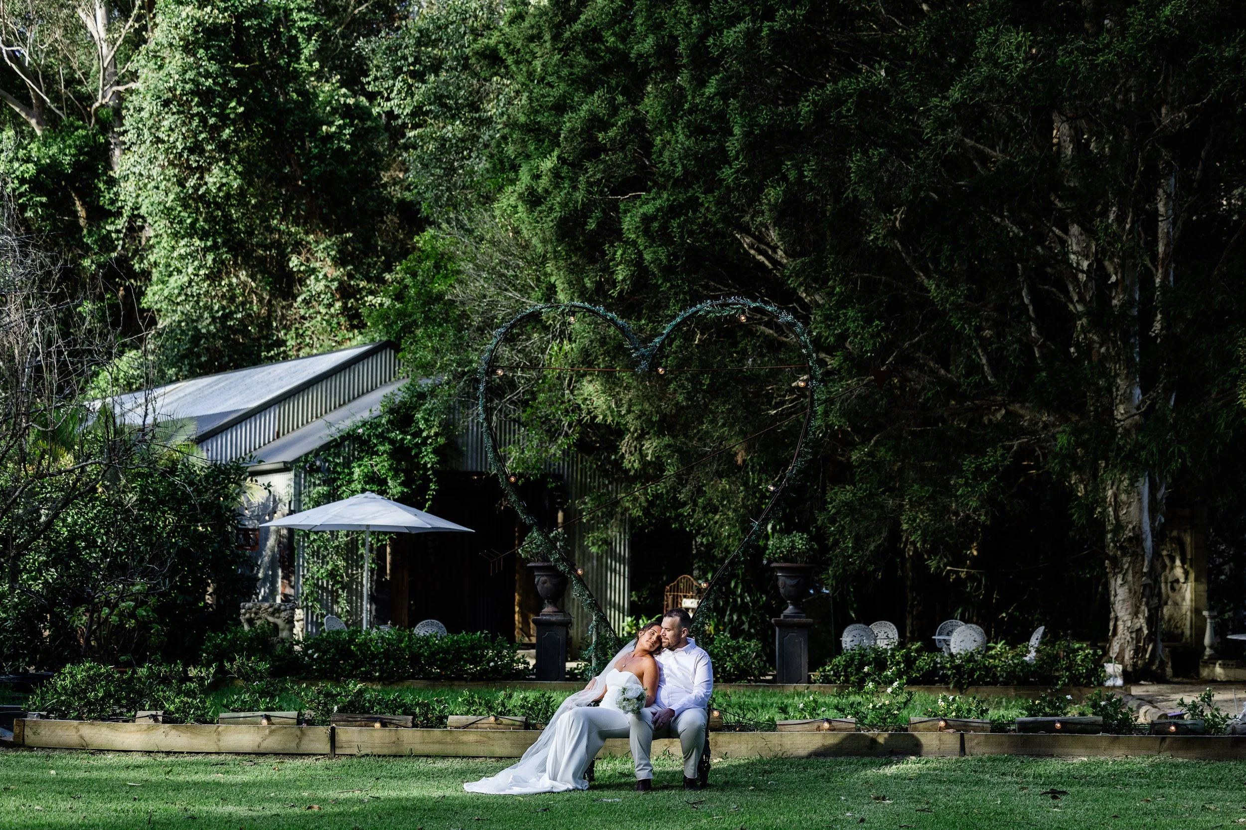 Bride and groom sitting on a bench in a garden, surrounded by trees, with a large heart-shaped decoration behind them, during their wedding.