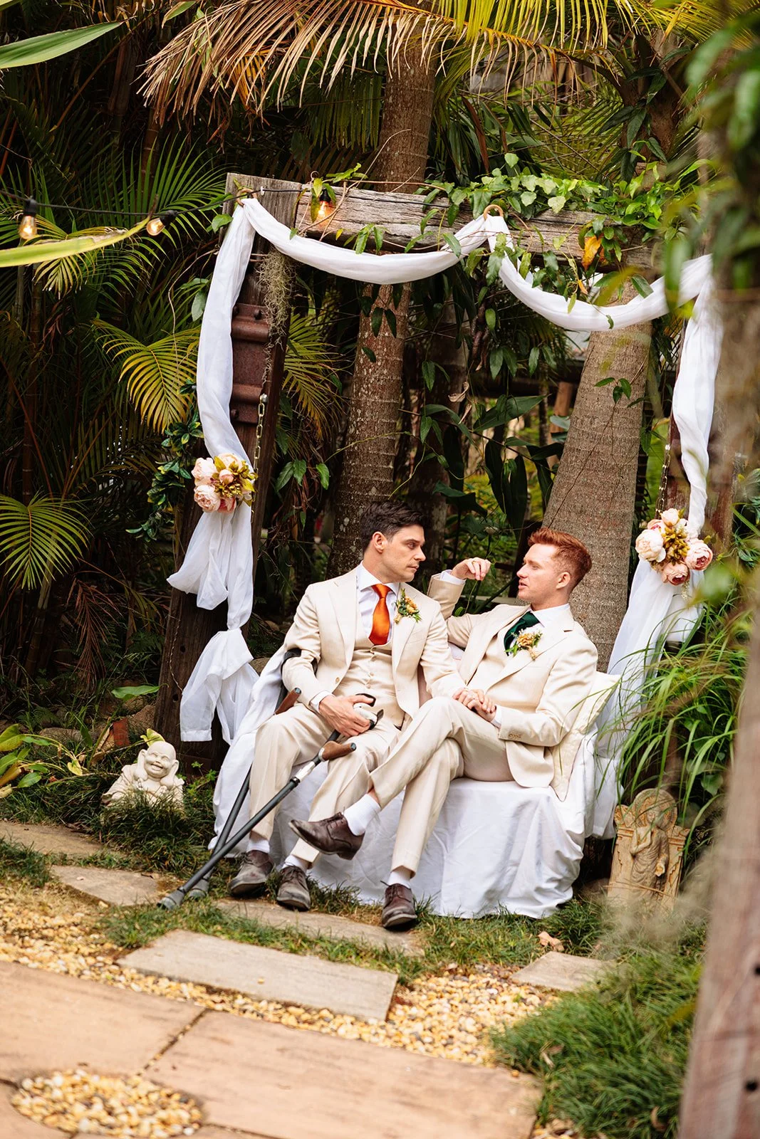 Two men in beige suits sitting on a white bench under a decorated archway with flowers and white fabric, surrounded by trees and lush greenery during an outdoor wedding ceremony.